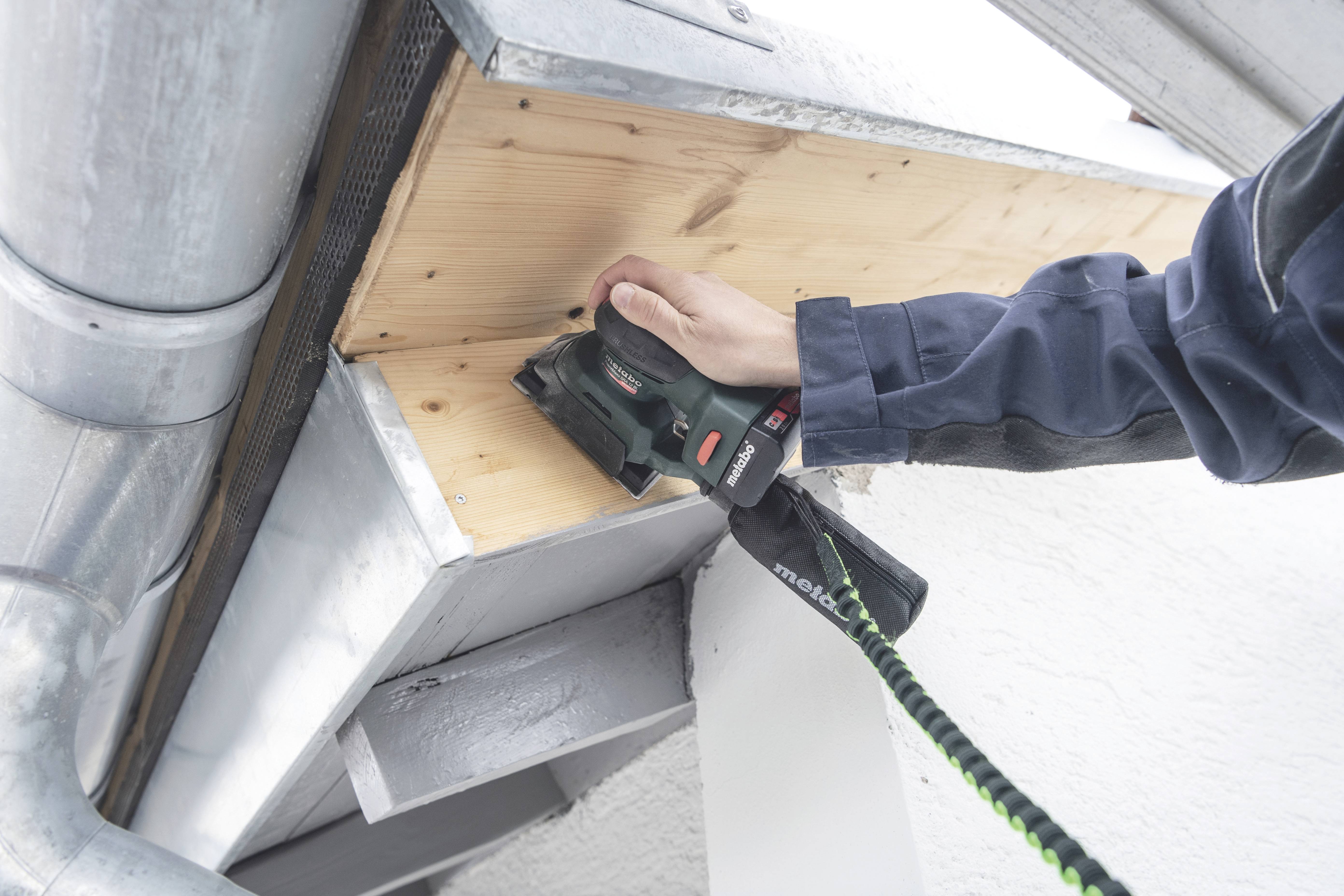 A person is sanding wood on an eaves overhang using an electric sander. Metal parts and drainage pipes are visible.