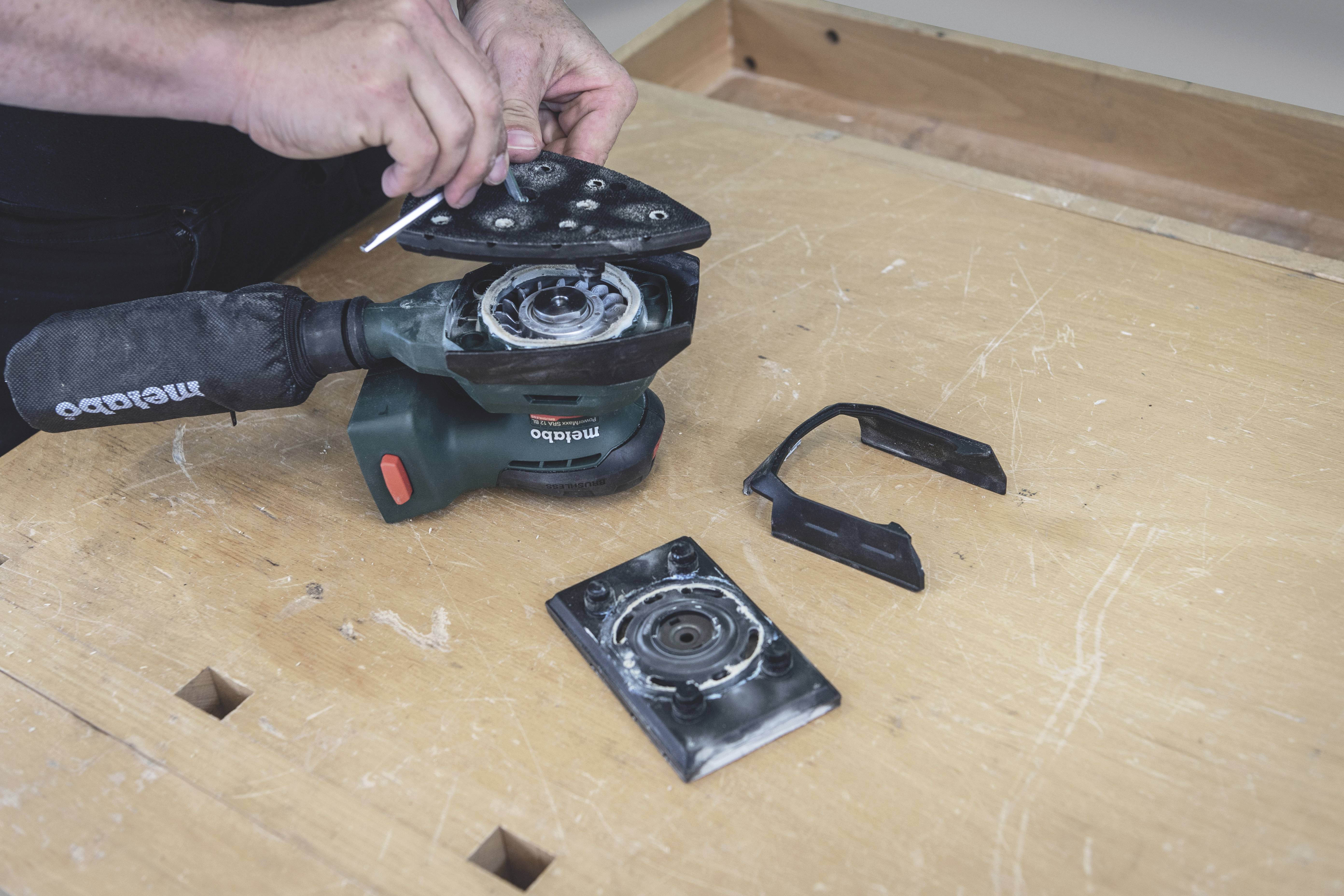 A person is repairing a green Metabo electric grinding machine on a wooden table. Tools and worn parts are lying beside it.