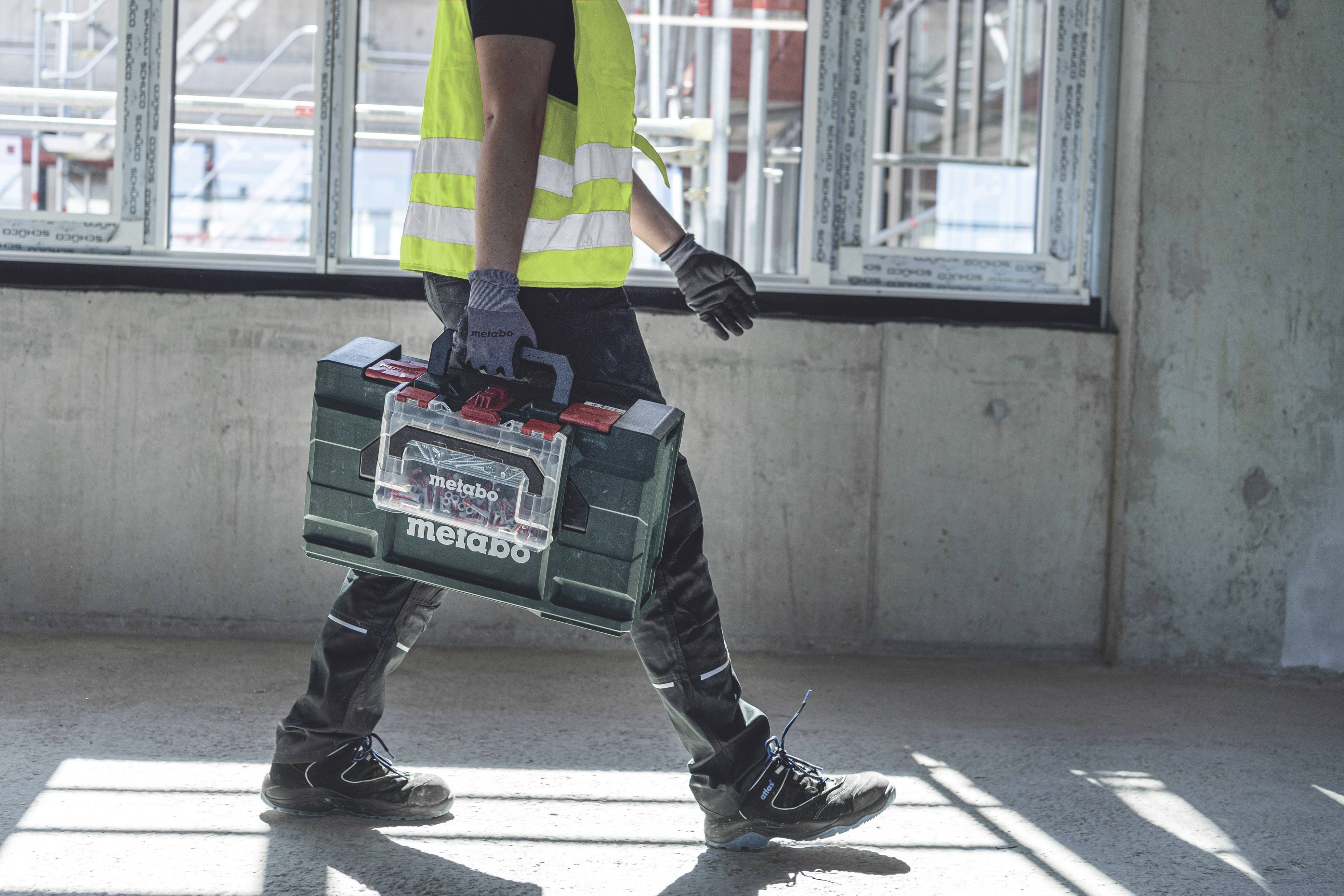 A construction worker in safety clothing is carrying a toolbox through a building site with large windows in the background.