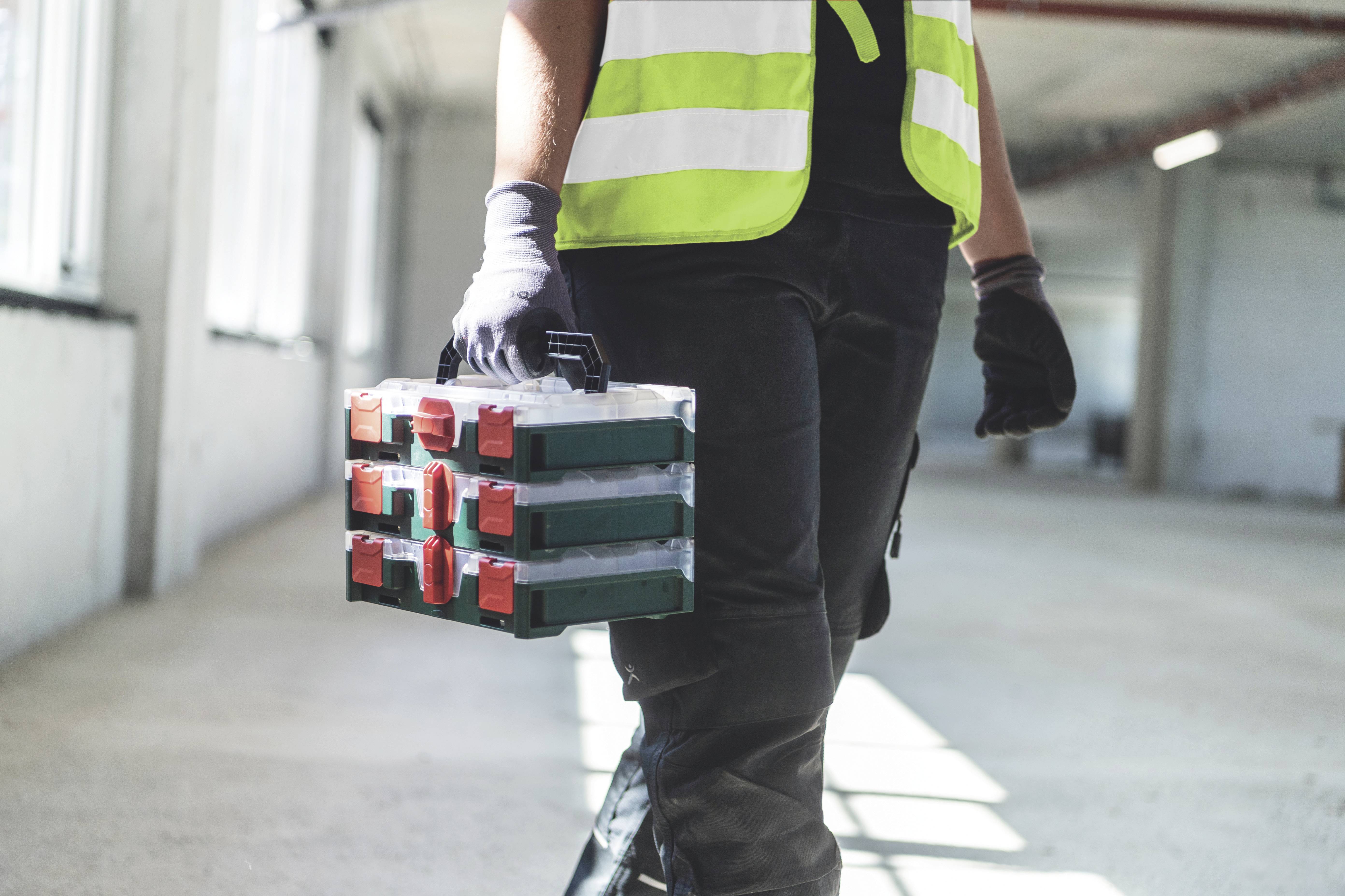 A person in work attire and a high-visibility safety vest is carrying a toolbox. They are in an empty, bright room.