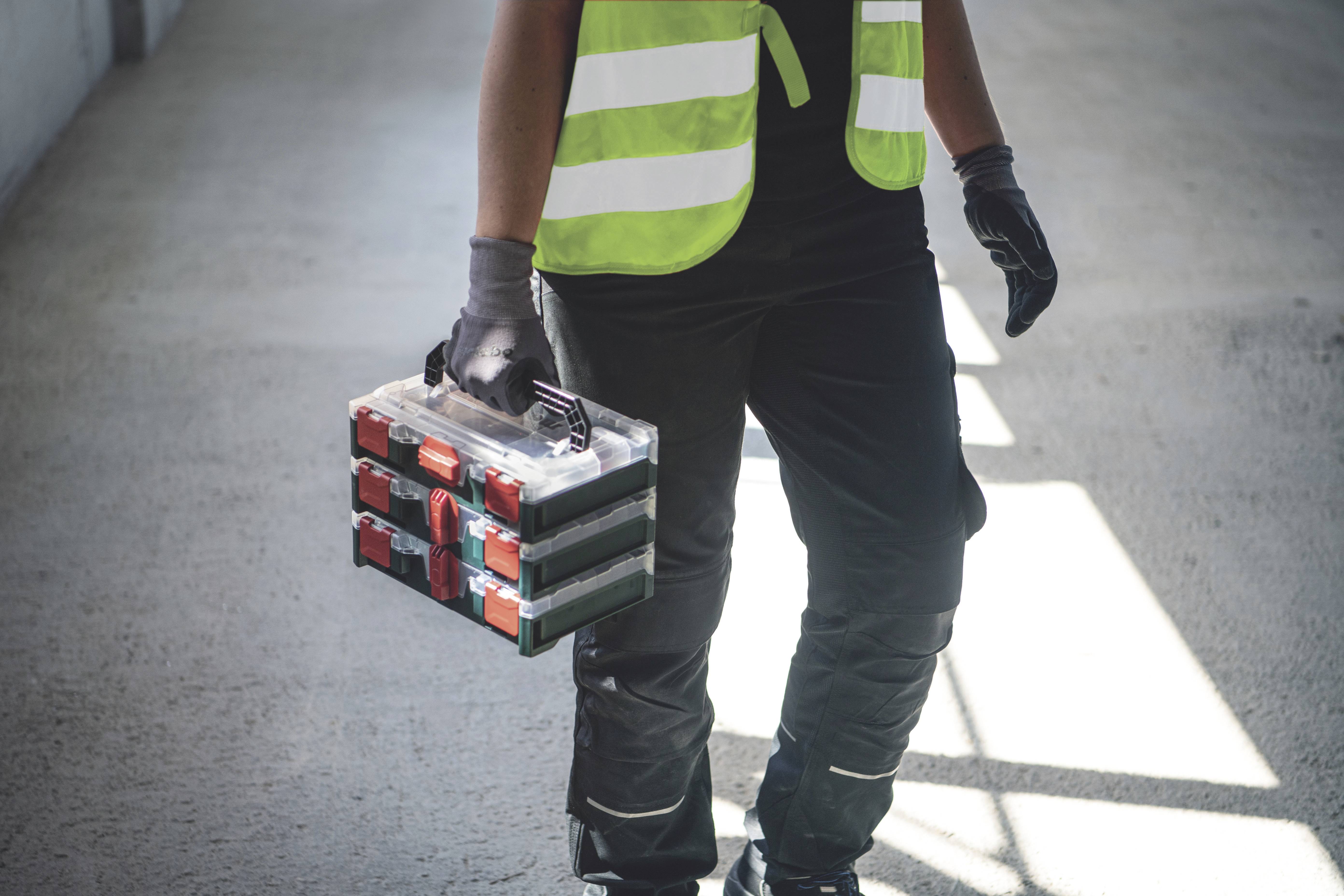 A person in workwear is carrying a toolbox in a light-filled environment.