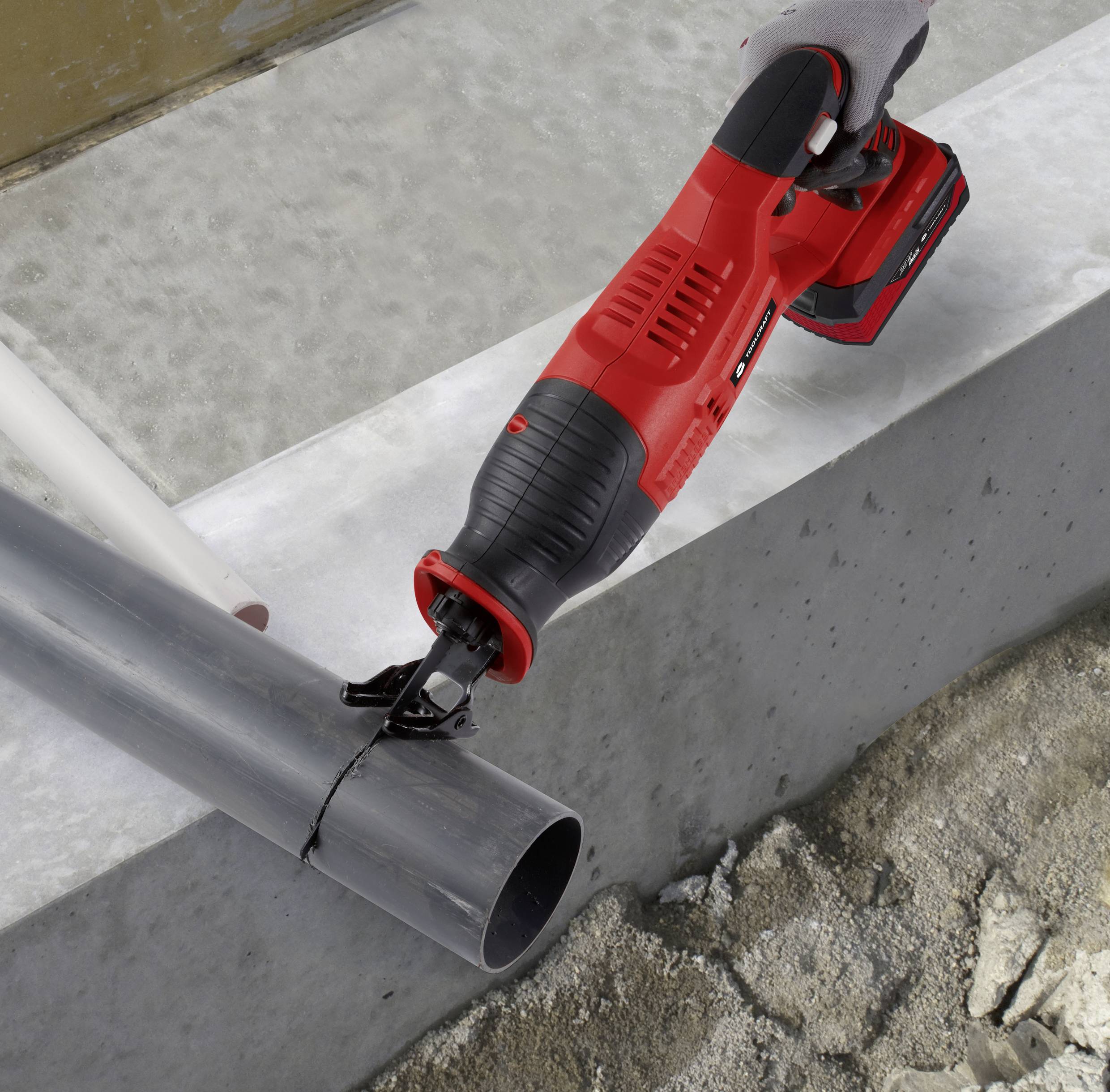 A red electric saw is being used to cut a grey plastic pipe on a concrete surface.