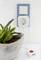 A plant pot is positioned next to a power socket. A white adapter with a red button is plugged into the socket.