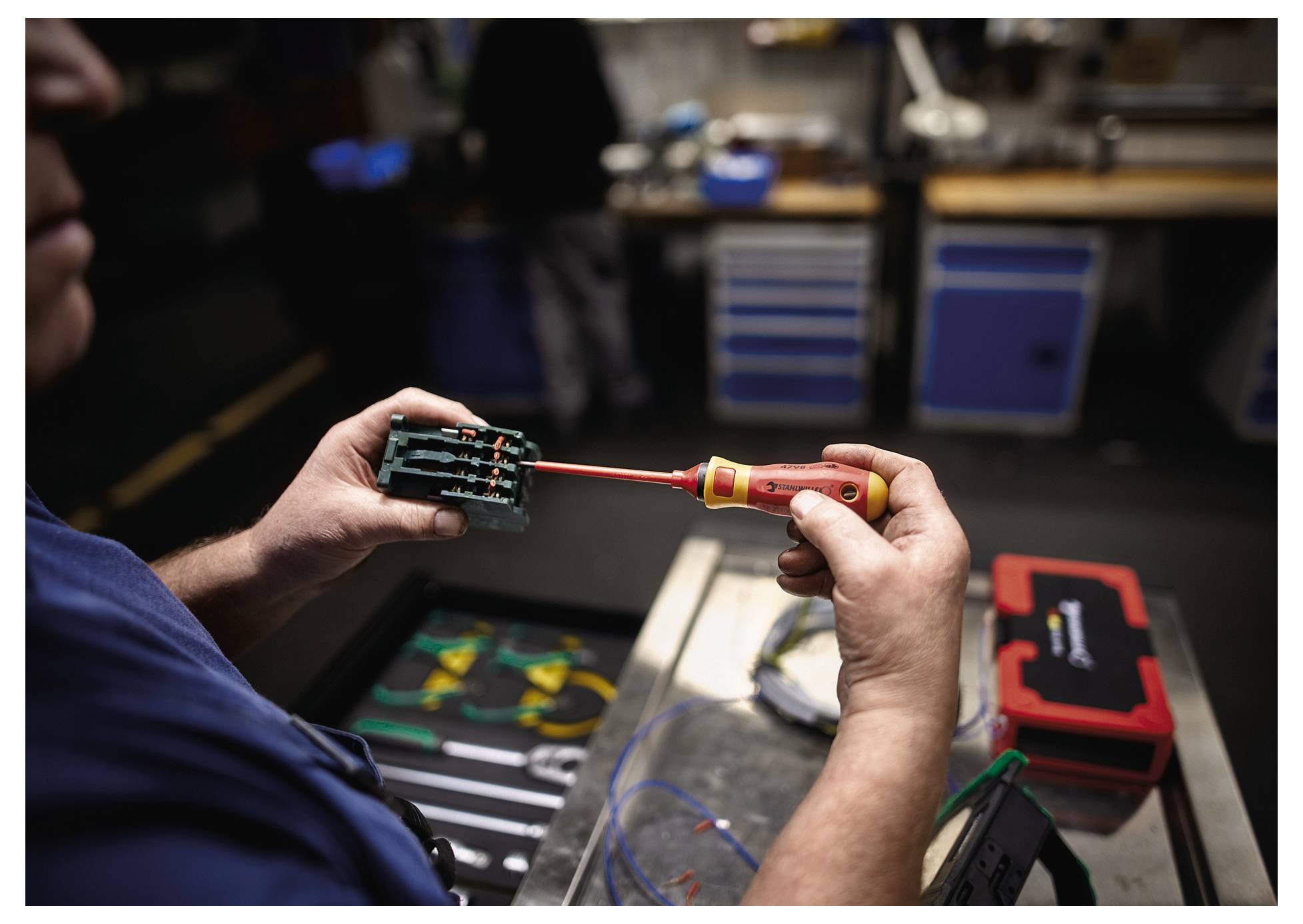 A person is using a screwdriver to work on a small circuit board in a workshop. Tools and equipment are visible in the background.