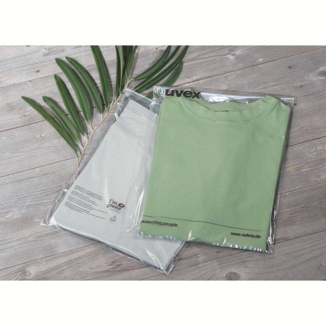 A green T-shirt and a white garment are lying in transparent packaging on a wooden floor. Green leaves are positioned beside them.