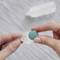 A hand holds a flat, round bottle cap; another hand colours it with a turquoise sponge.
