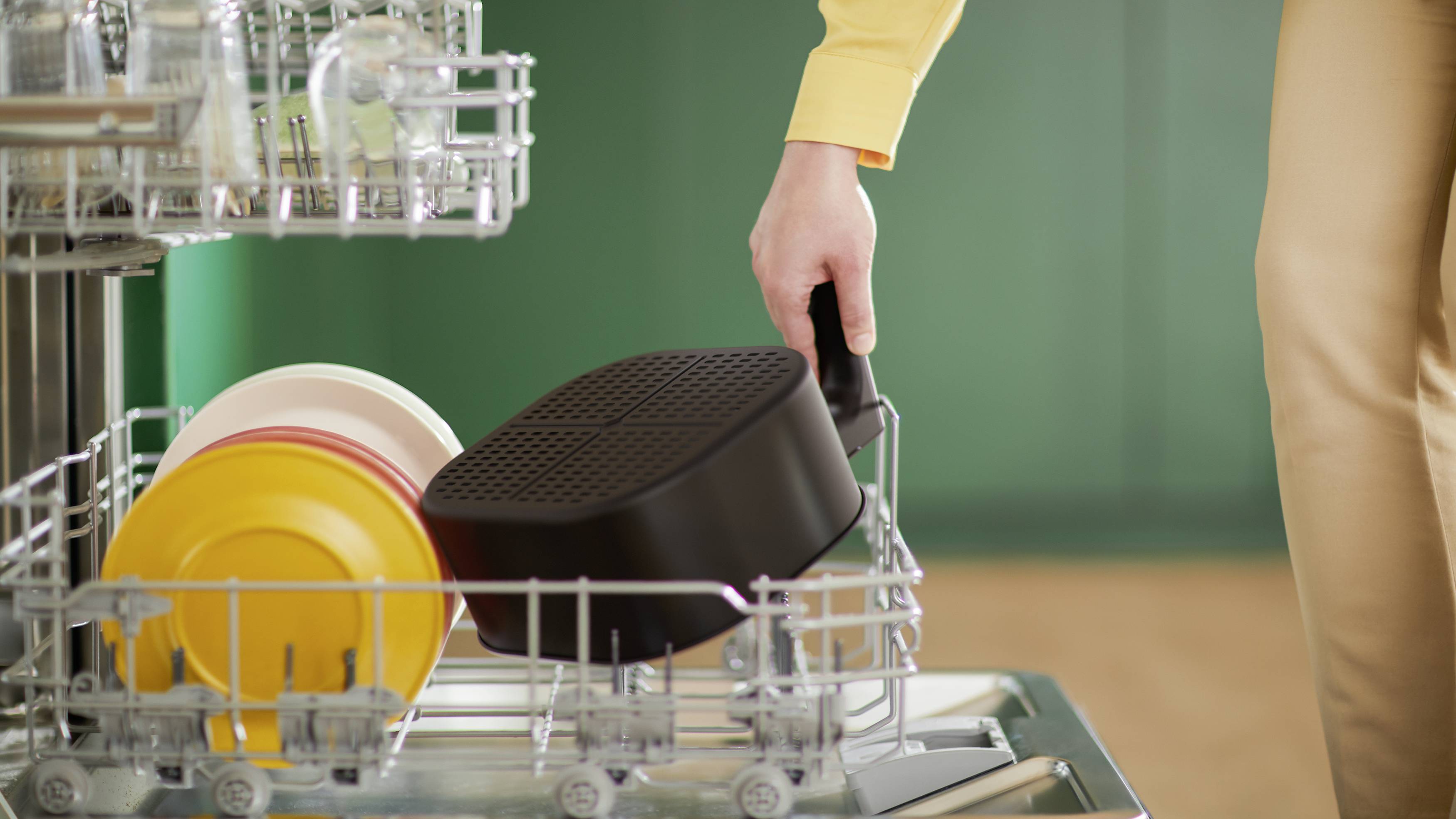 A person is loading a pan into the dishwasher. The top rack is filled with glasses, and the bottom section contains colourful plates.