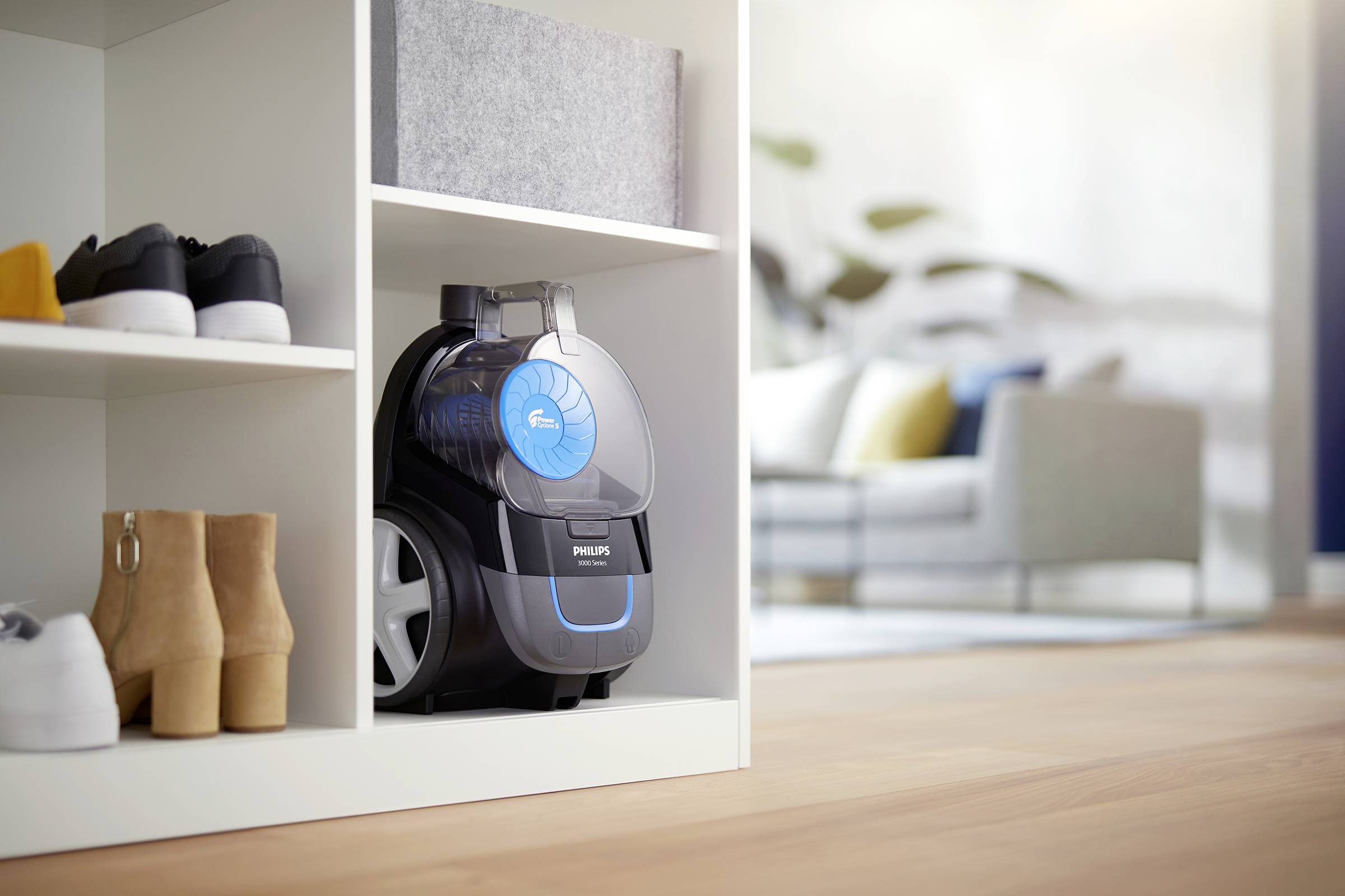 A compact, black vacuum cleaner with blue accents is placed on a white shelf next to shoes and boxes, with a living room in the background.