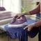 A person is ironing a garment on an ironing board. A purple iron is being used. Blurred furniture can be seen in the background.