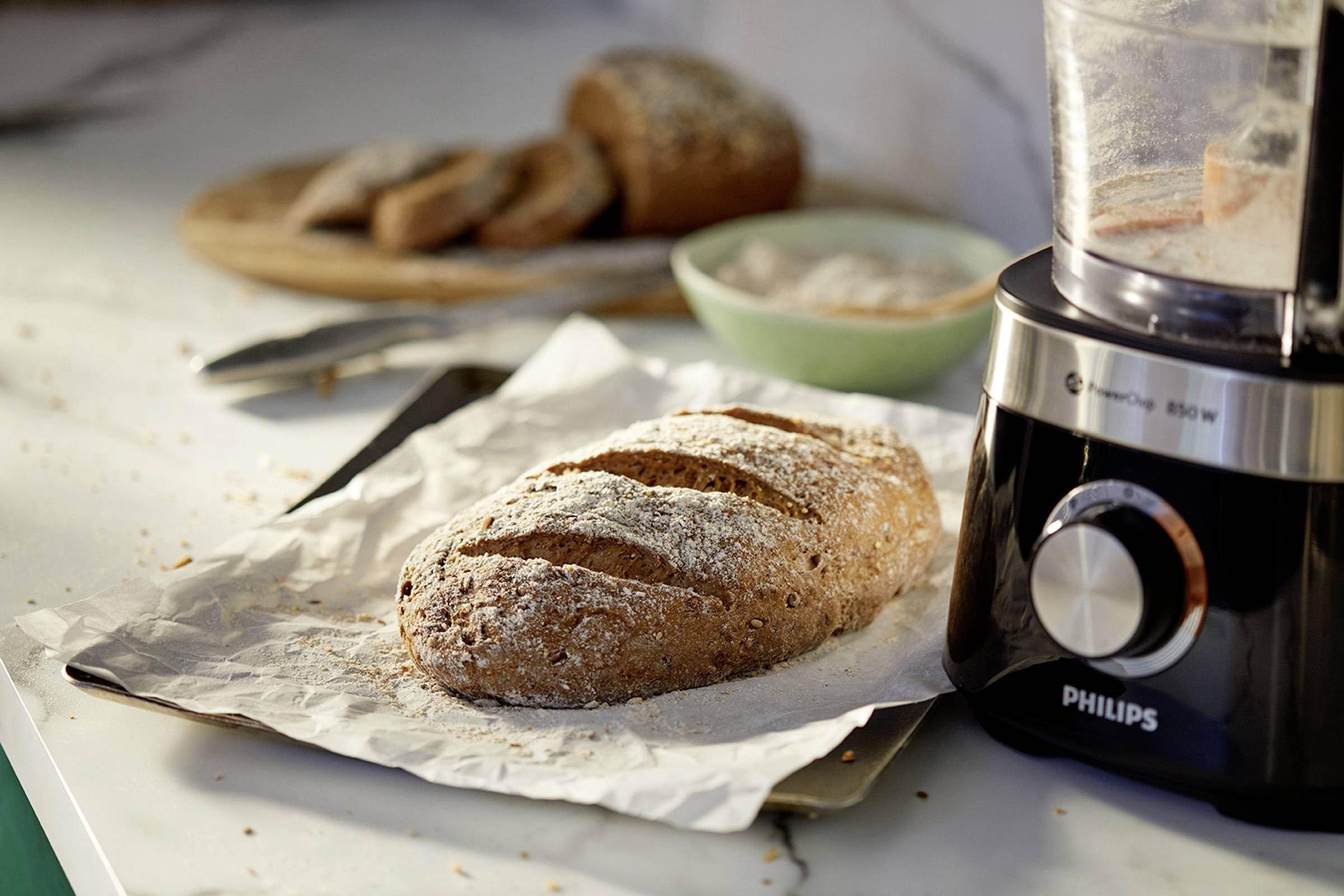 A freshly baked loaf of bread sits on baking parchment on a table, next to a food mixer. In the background, additional slices of bread can be seen.