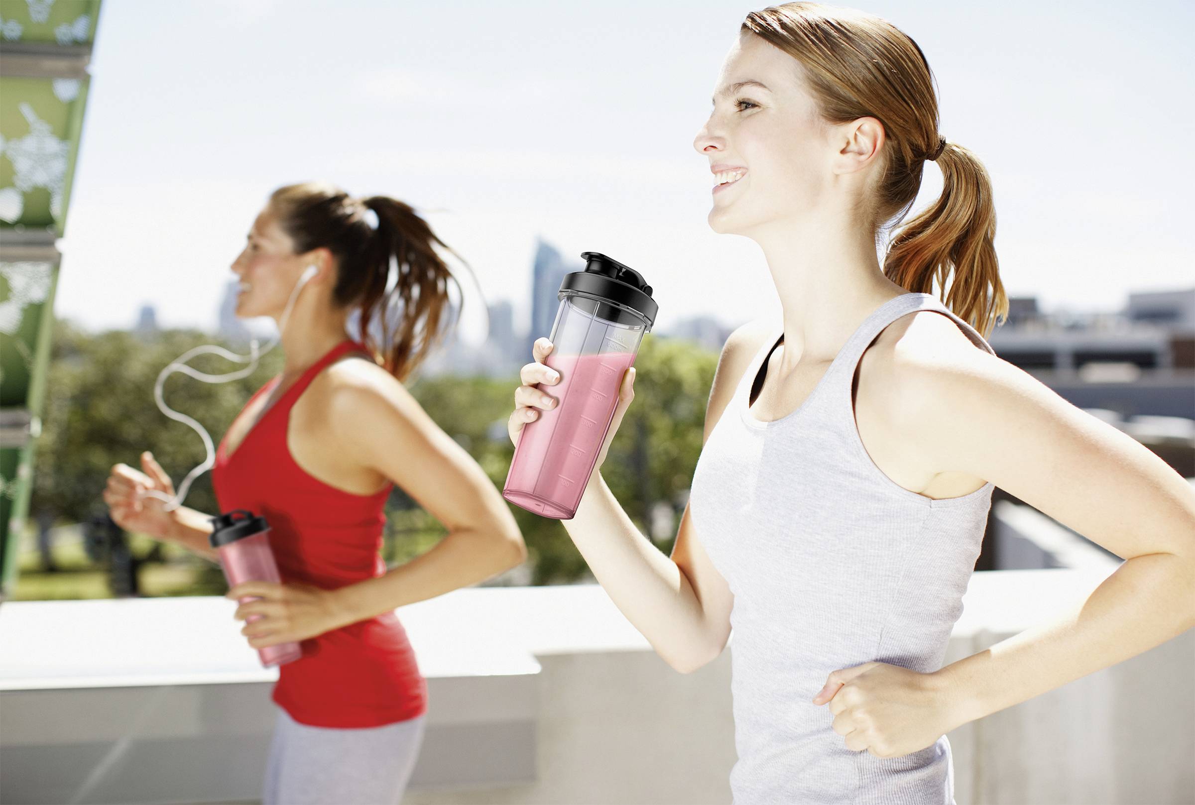 Two women are jogging in sunny weather and holding water bottles. One woman is listening to music through headphones. Trees can be seen in the background.