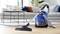 A blue vacuum cleaner is switched on, standing on a wooden floor in a living room with a grey sofa and coffee table in the background.