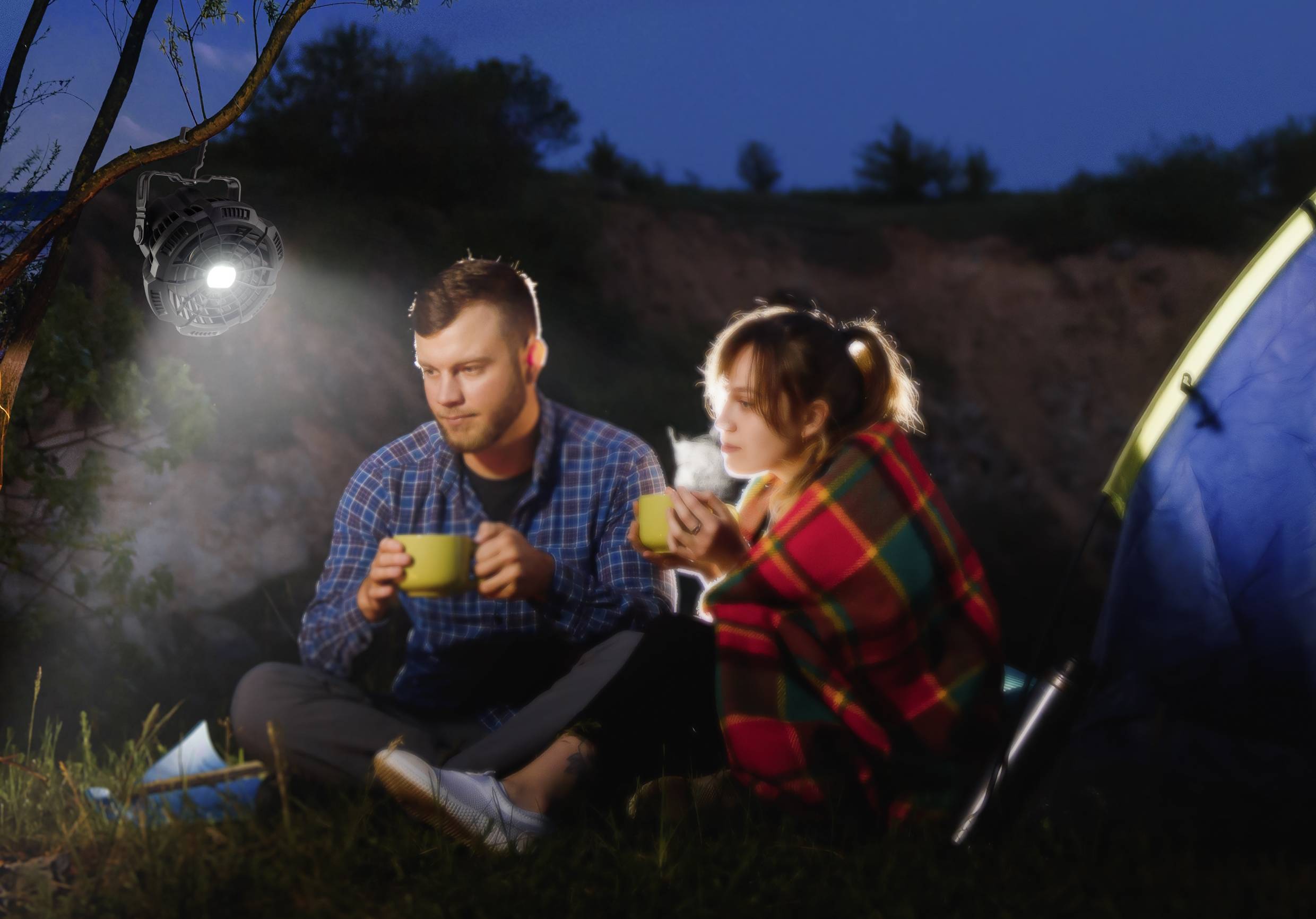 A man and a woman are sitting outdoors on a meadow at night. They are holding mugs and wrapped in blankets, next to a tent.
