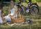 Two people are sitting on a blanket in the park with a picnic hamper. Bicycles are standing in the background.