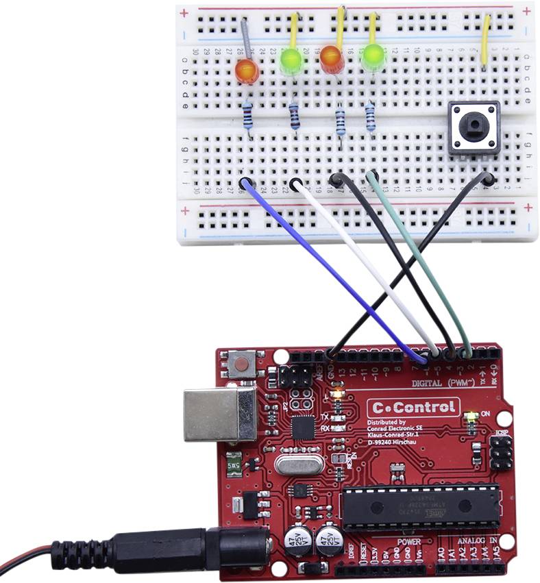 A red microcontroller board is connected via cables to a breadboard. On the breadboard, there are four LEDs, a push button, and resistors.