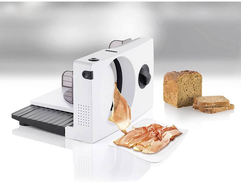 A white electric bread slicer is cutting thin slices of ham; two slices of bread are lying beside it.