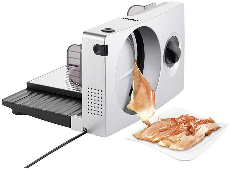 An electric slicing machine cuts thin slices of ham, which are lying on a white plate.
