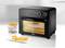 Black mini oven on white background, showing LED display '230C 00:45'. Filled baking dish inside, with a plate of mini pizzas and a deep-fat fryer basket beside it.