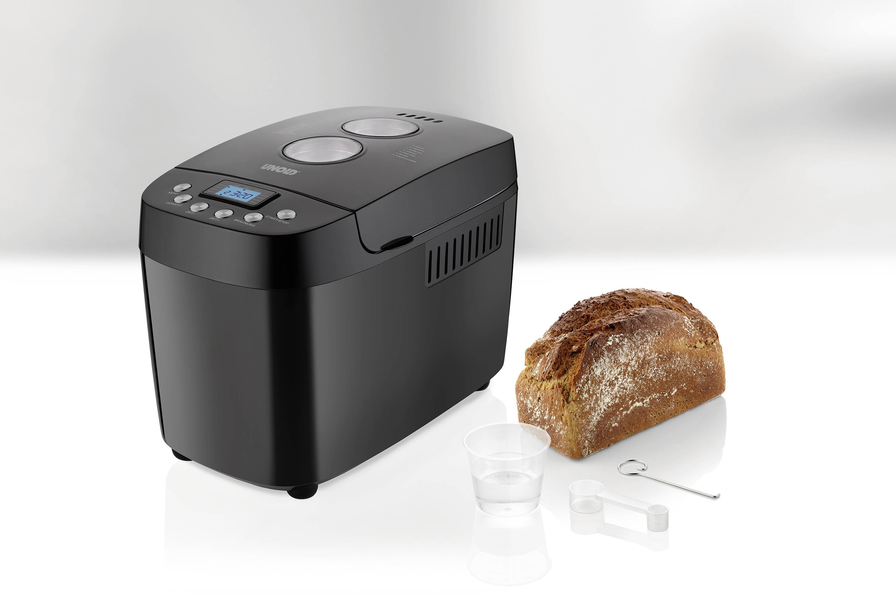 Black bread maker on a kitchen counter with a freshly baked loaf beside it. Accessories including measuring cup and hook.