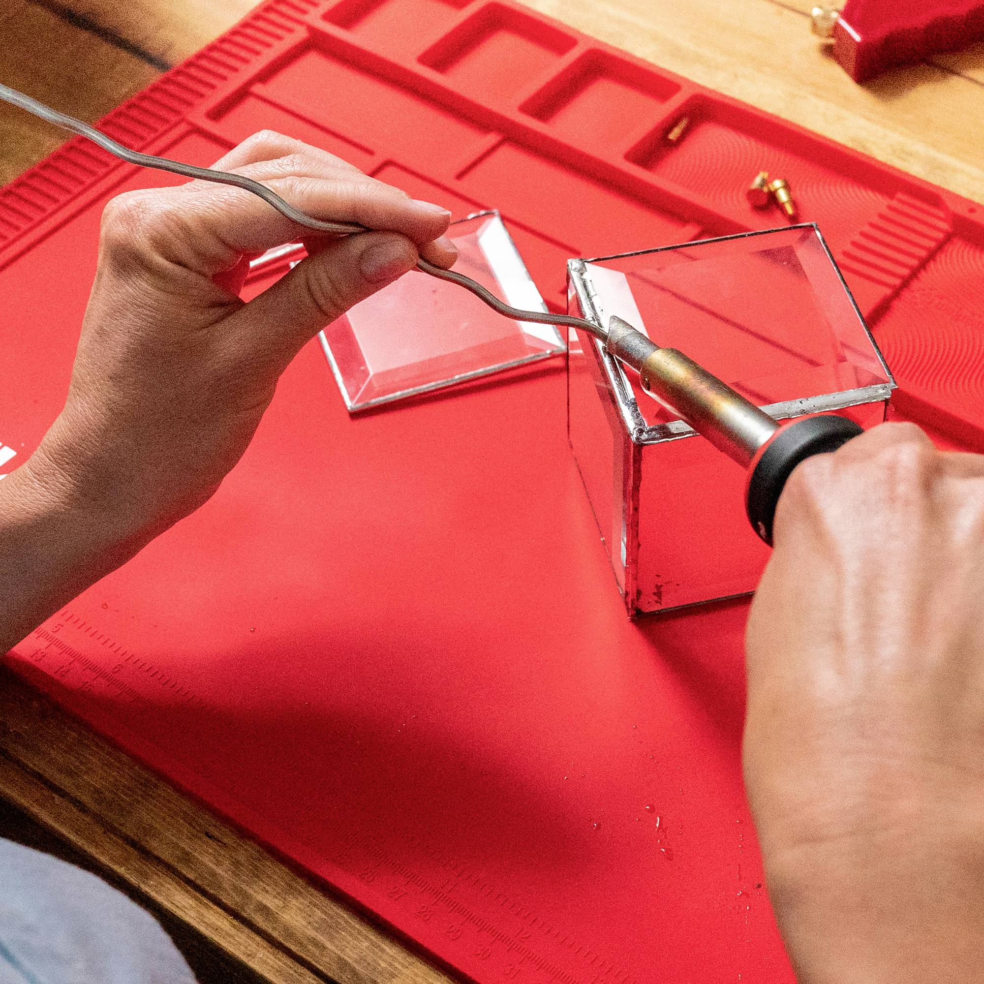 Hand holding soldering iron, soldering onto a glass cube on a red surface. Two hands visible, focused work scene.