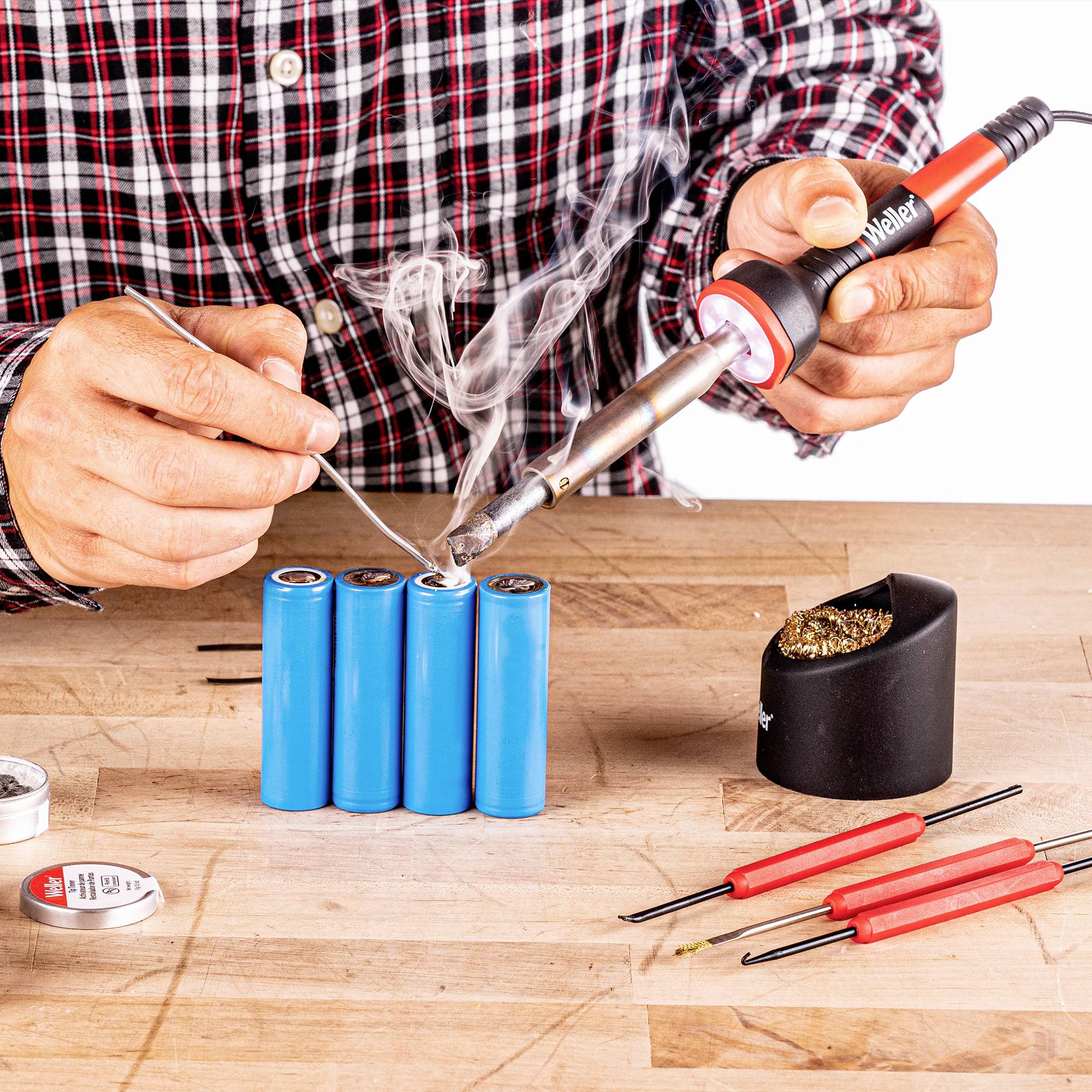 A person is soldering several cylindrical, blue batteries with a soldering iron. Smoke rises, and tools are laid out ready.