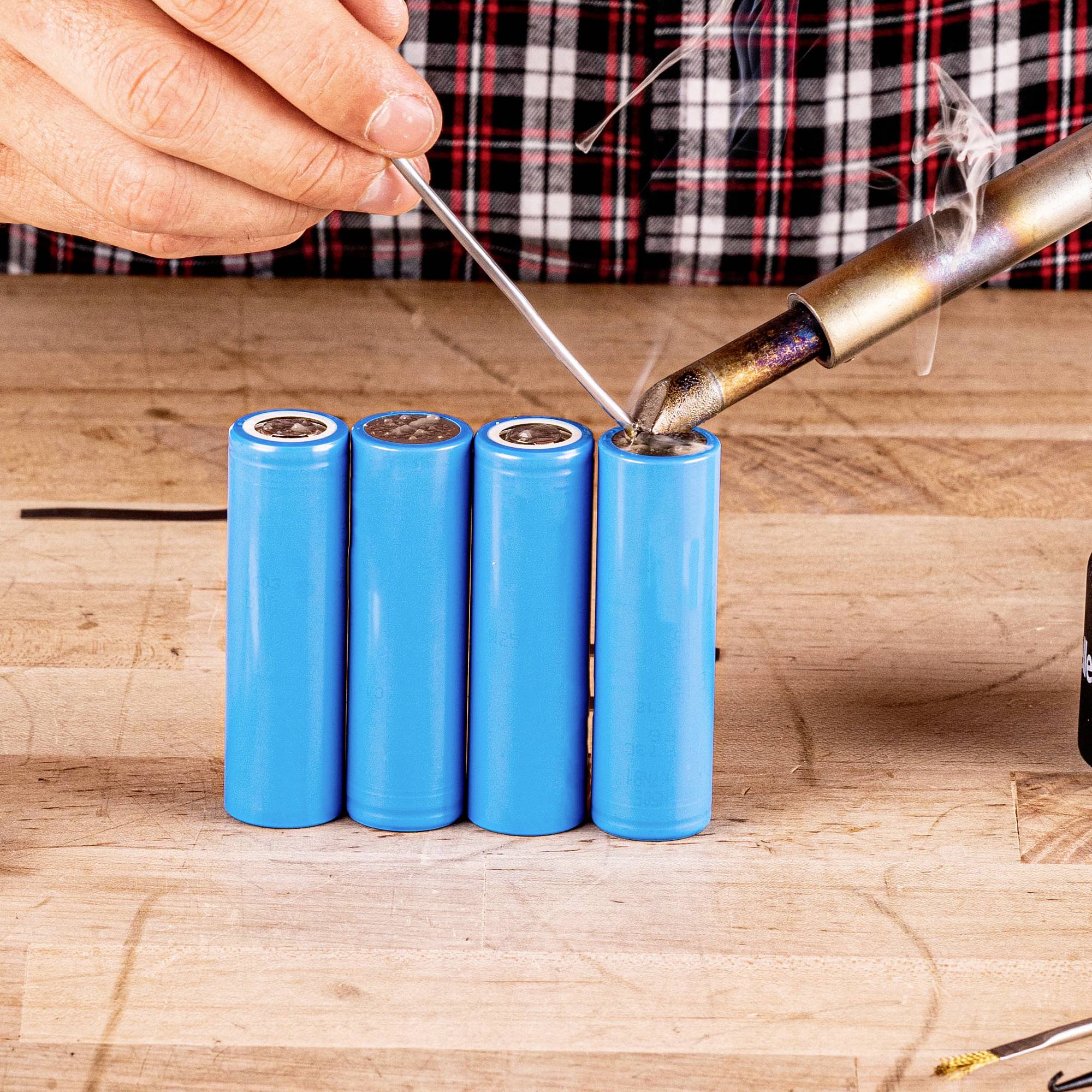 A soldering iron touches the top of a blue battery, while a person holds the two objects together to connect them.