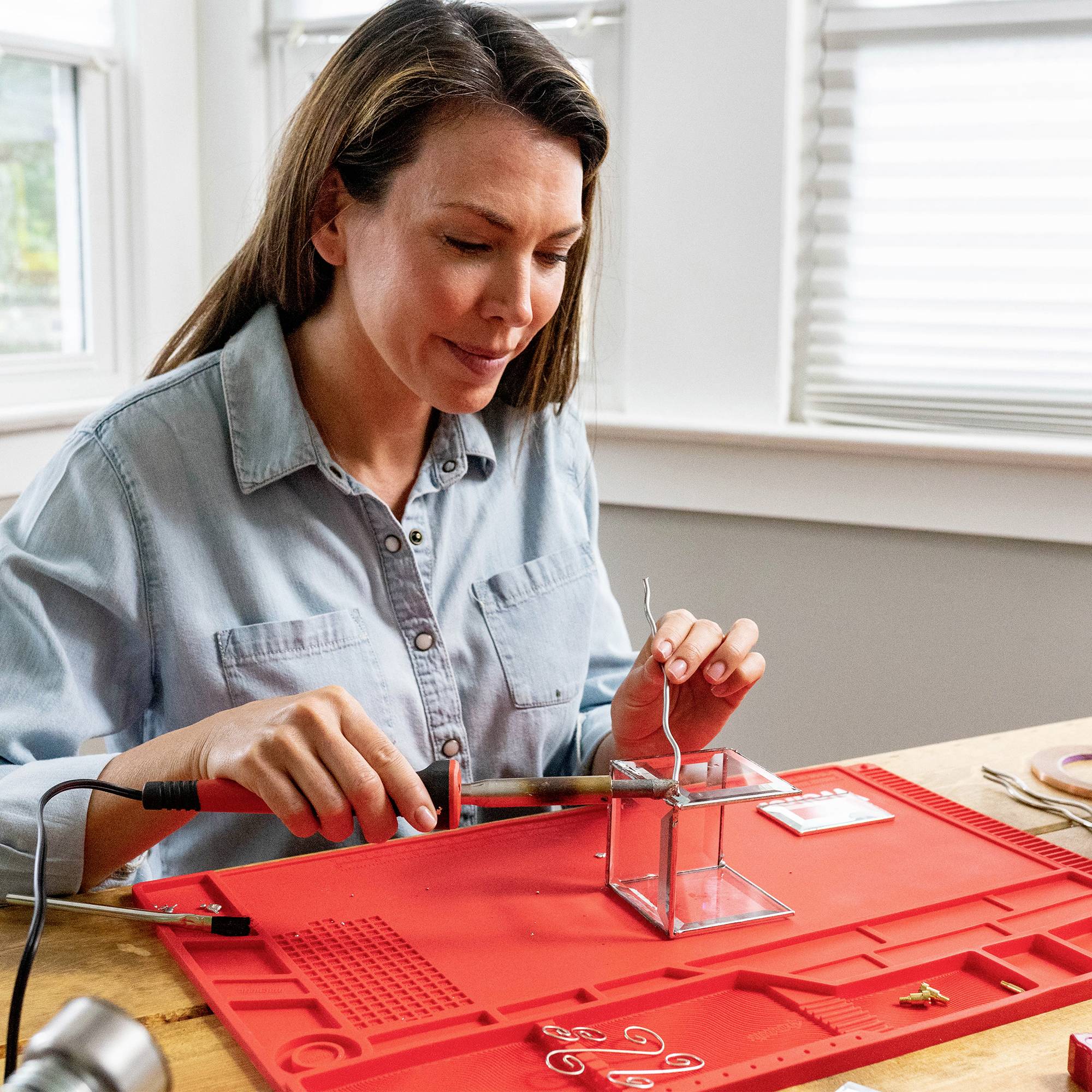 A woman is soldering on a red workboard in a bright room, surrounded by tools and accessories.
