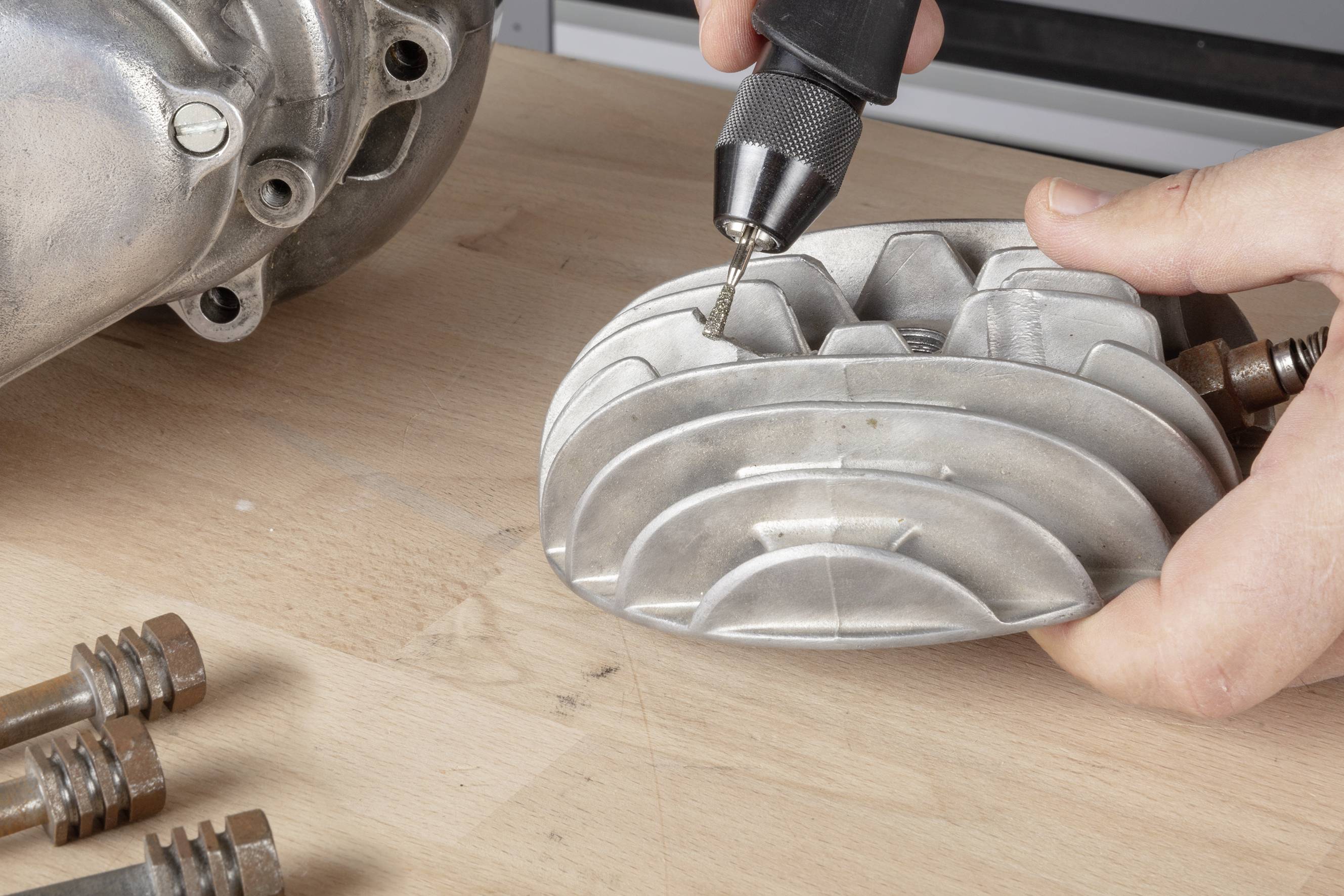 A person is drilling a metallic machine part on a table. Further parts are visible in the background.