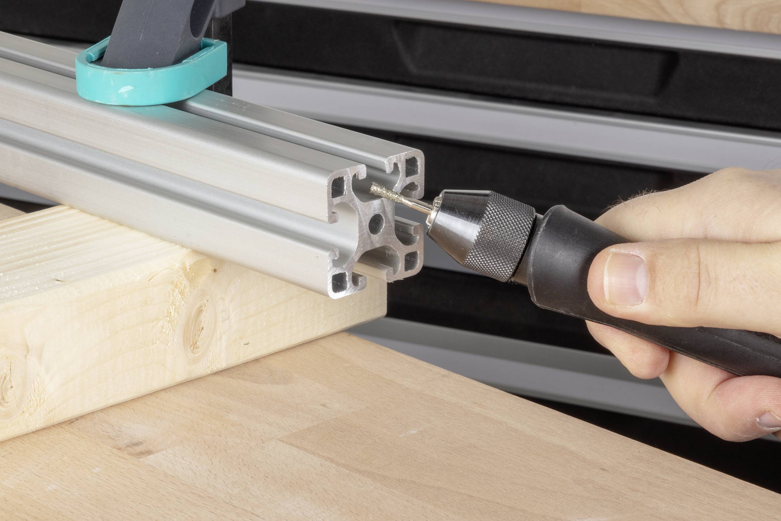 A person is drilling a hole in an aluminium profile using a cordless drill, with the profile resting on a wooden block.