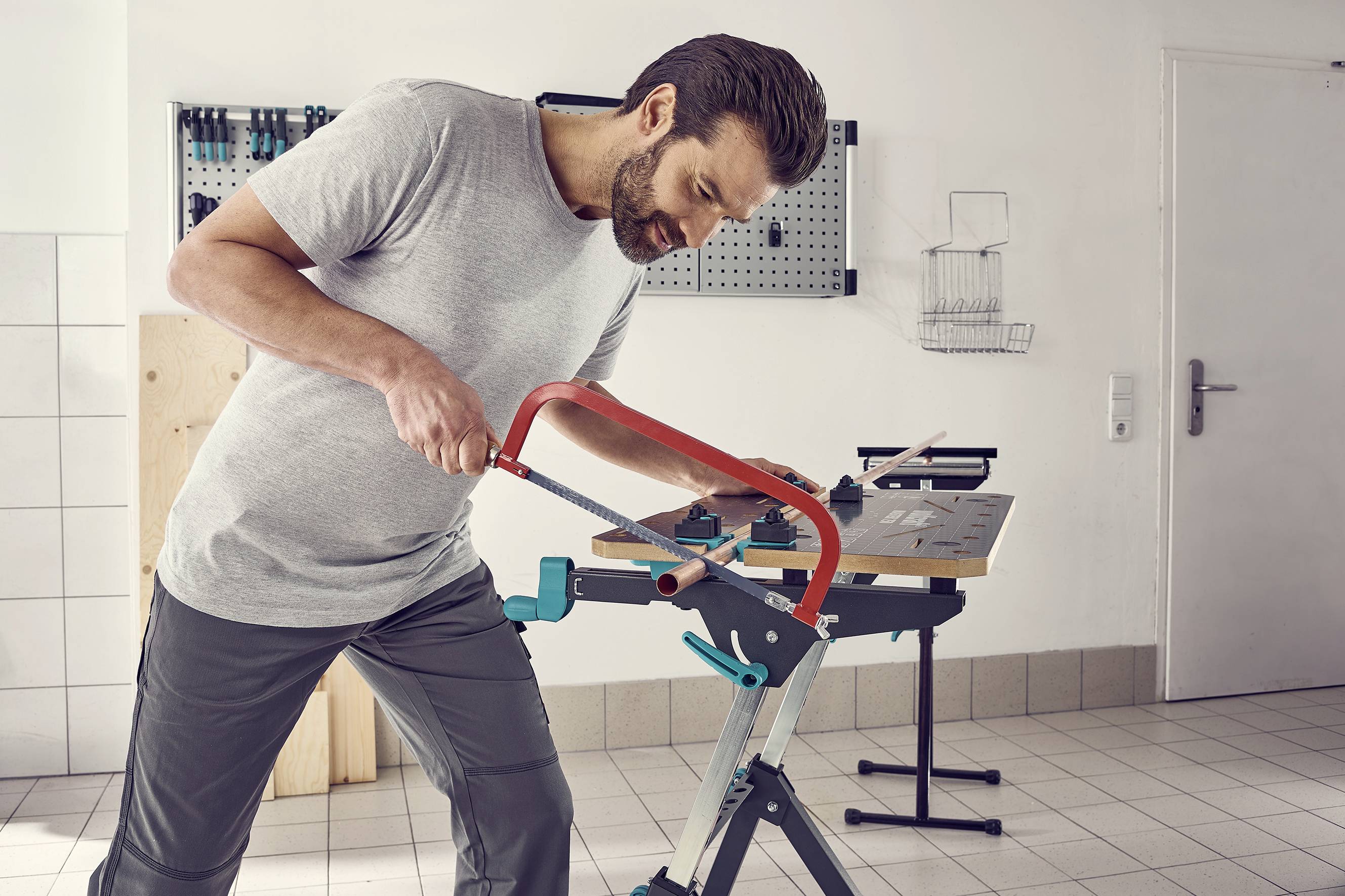 A person sawing a piece of wood in a workshop. Background: Tools hanging on the wall, bright light, tiled floor.