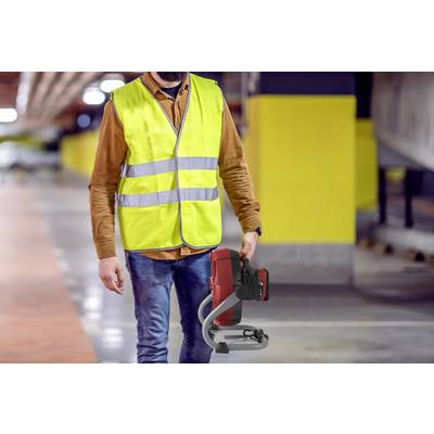 A person wearing a yellow high-visibility vest is walking through a car park and carrying a red electric tool. The surroundings are dimly lit.