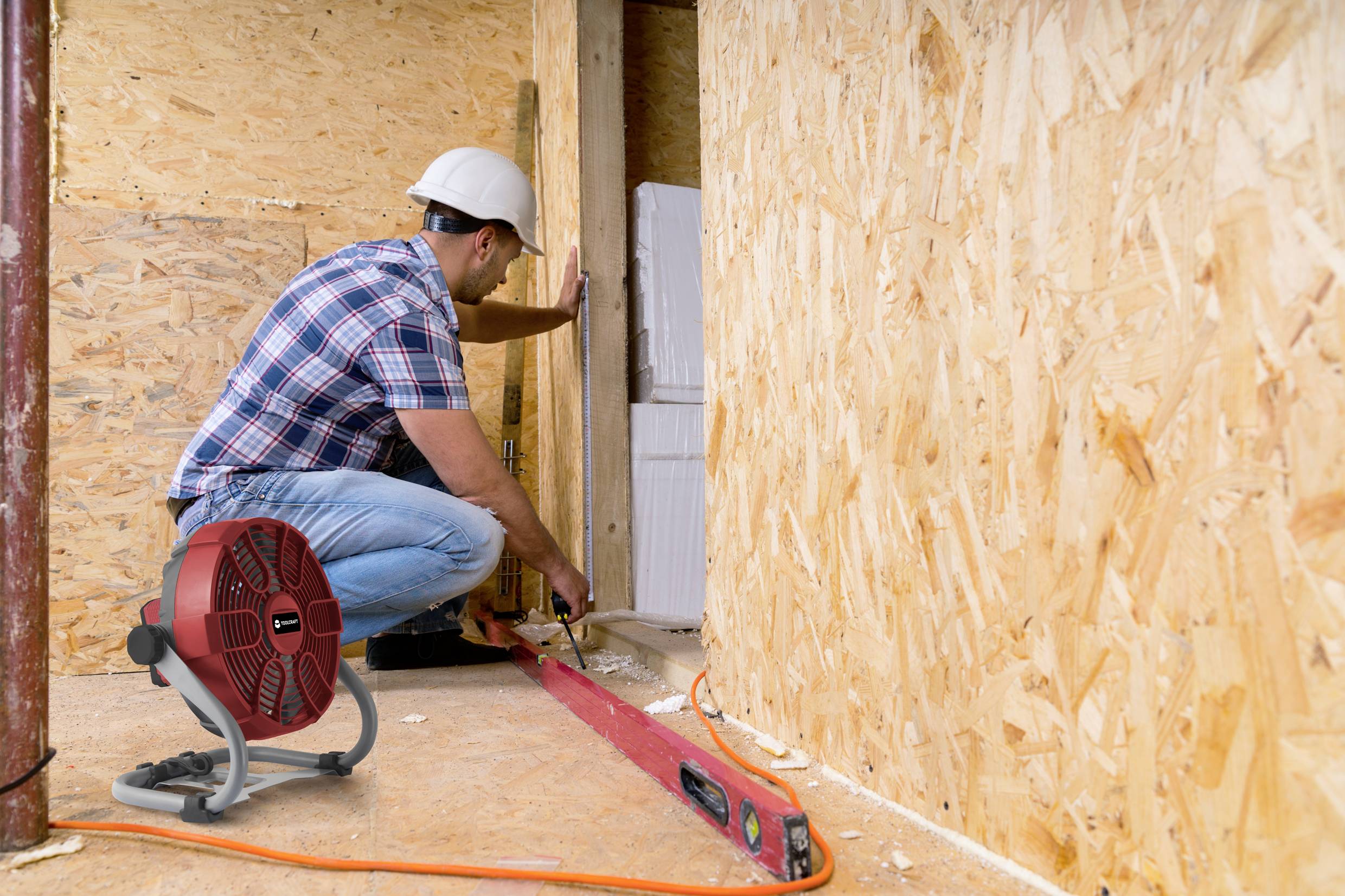 A person wearing a hard hat is working in a wooden room with a tape measure and spirit level. A red heater is standing on the floor.