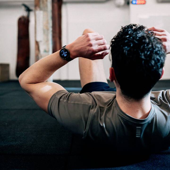A person is doing sit-ups on a mat in a gym. They are wearing a dark grey t-shirt and a wristwatch. Sports equipment can be seen in the background.