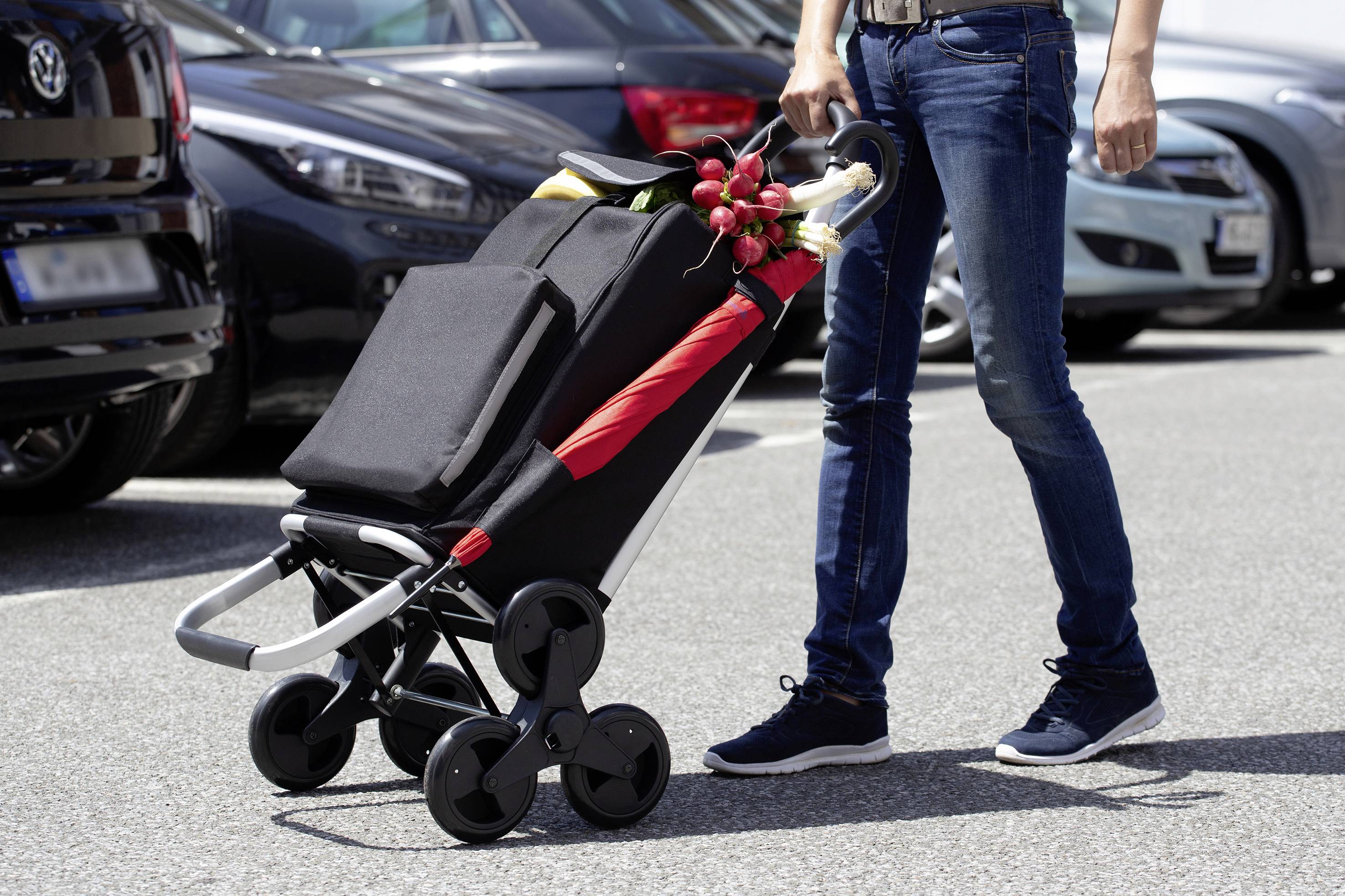 'A person wearing jeans and trainers is pushing a loaded shopping trolley across a car park, with cars in the background.'