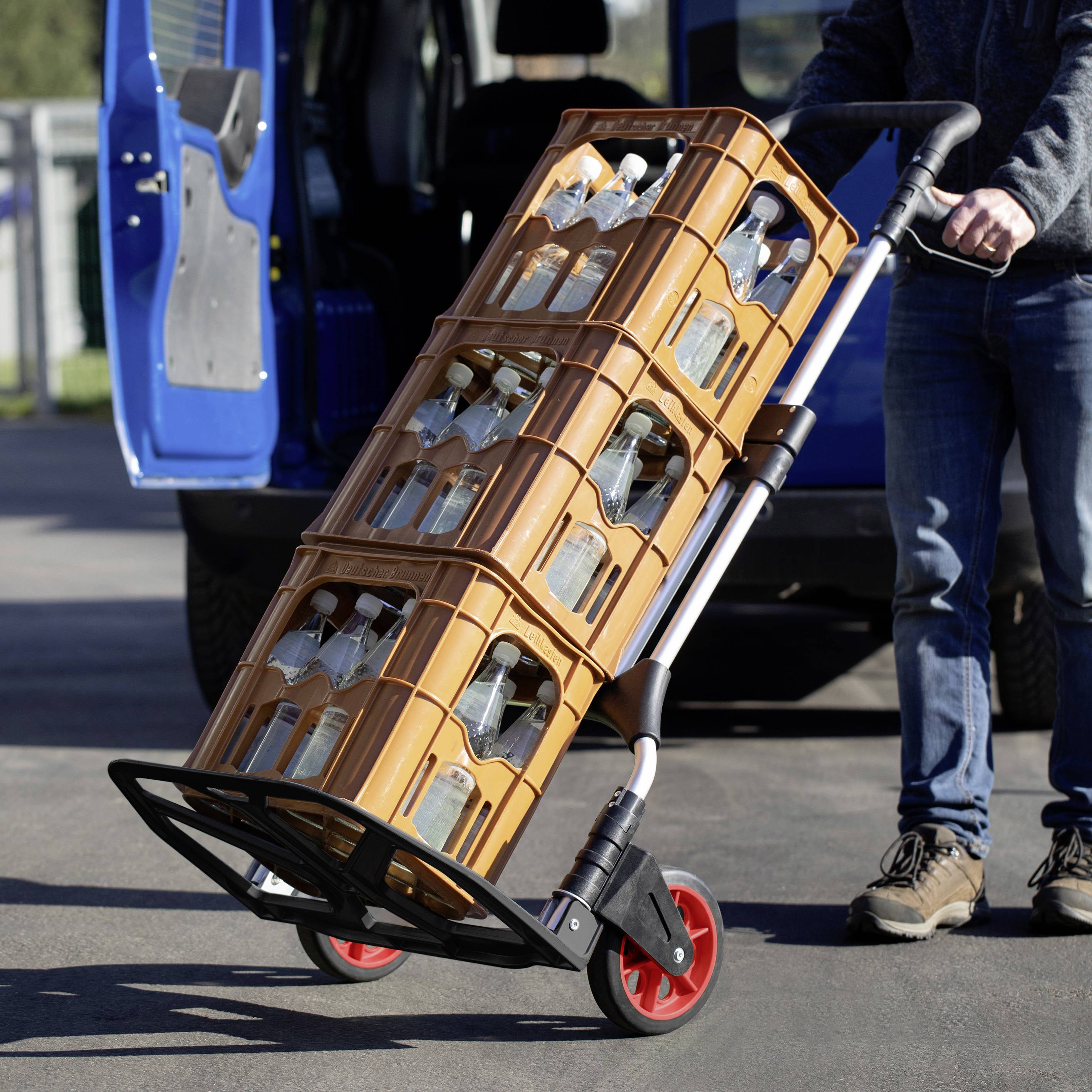 A person is transporting several crates of glass bottles on a sack truck in front of an open delivery van.
