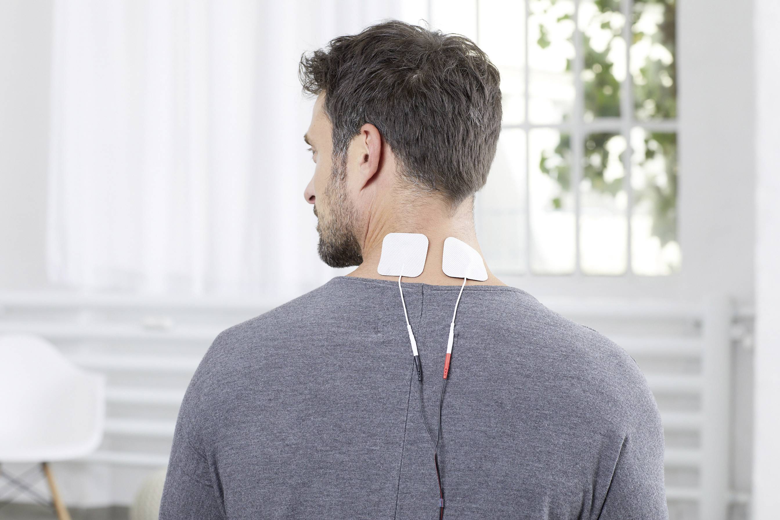 A man sits with his back to the camera. Two electrodes are attached to his neck, connected by cables, in a bright room.