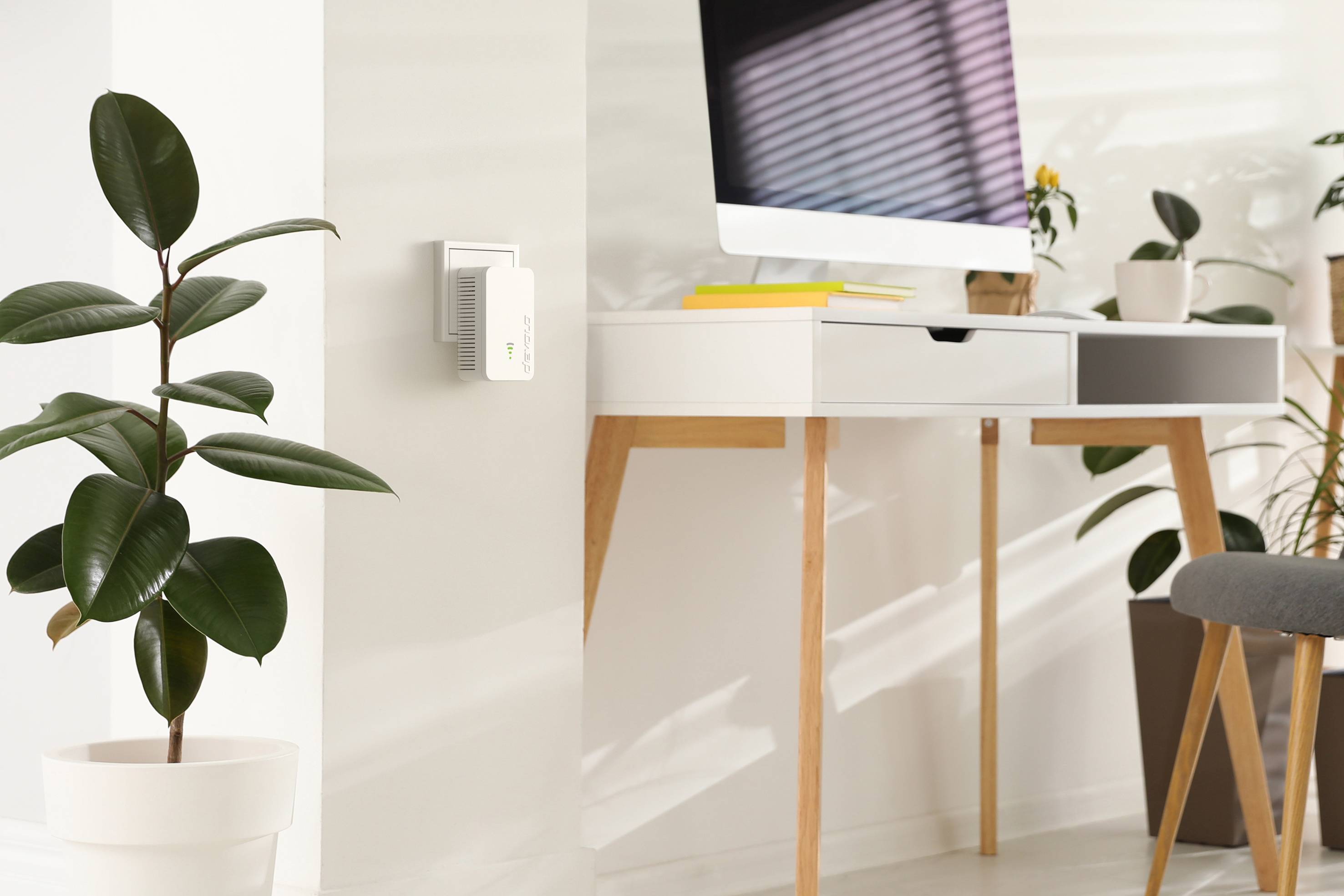 Modern workspace with a light wooden desk, computer and houseplants. A device on the wall displays a green light.