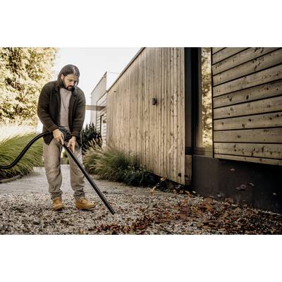 A man is using a leaf blower to clear autumn leaves from a gravel path beside a modern wooden house.