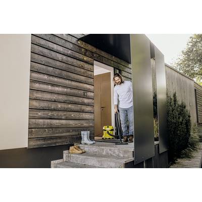 A person is standing on a house step and cleaning the doormat with a vacuum cleaner. Wellington boots and a pair of shoes are on the steps.