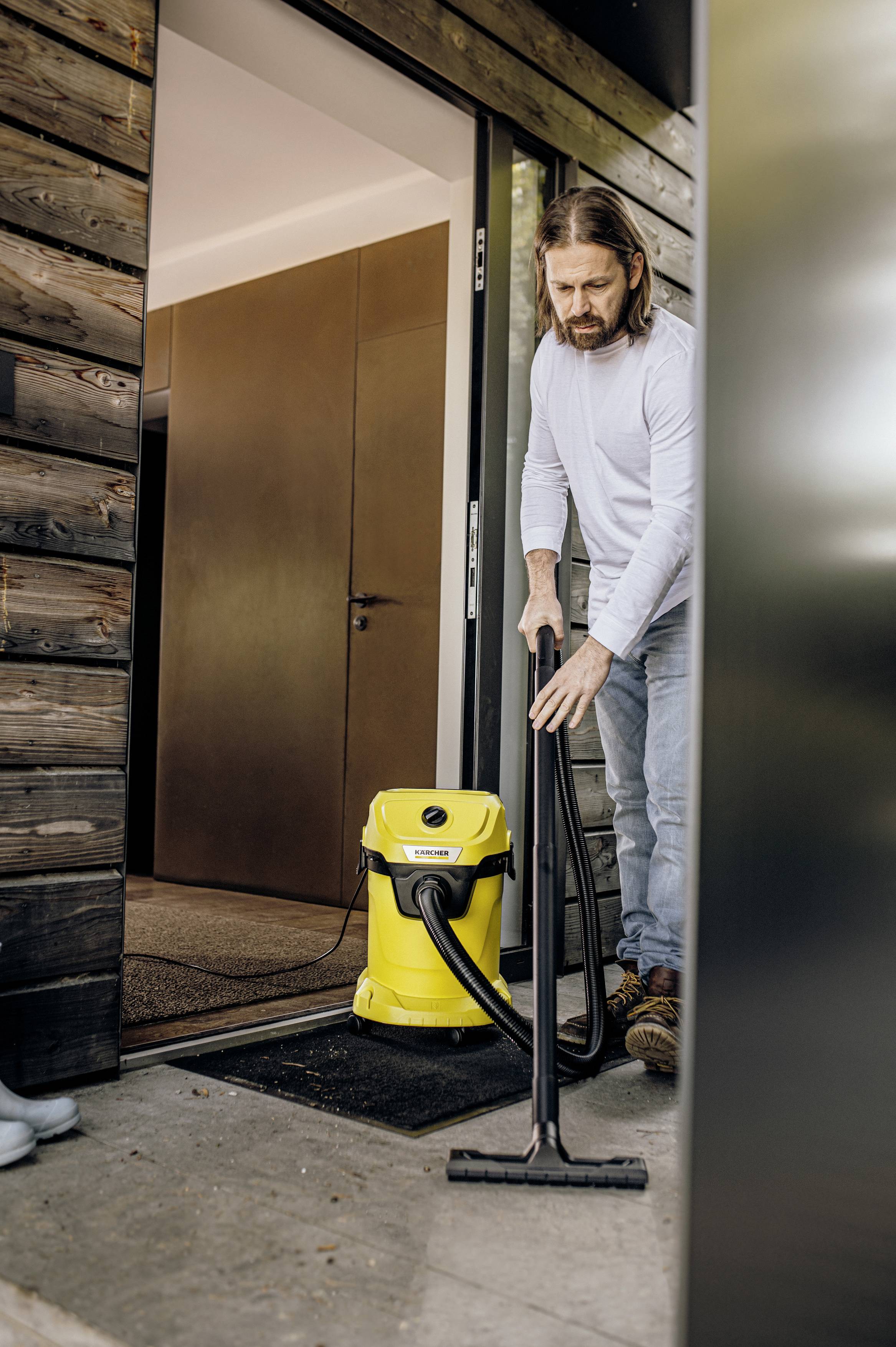 A man is cleaning the floor with a yellow vacuum cleaner while standing in front of a door. He is wearing a light top and jeans.