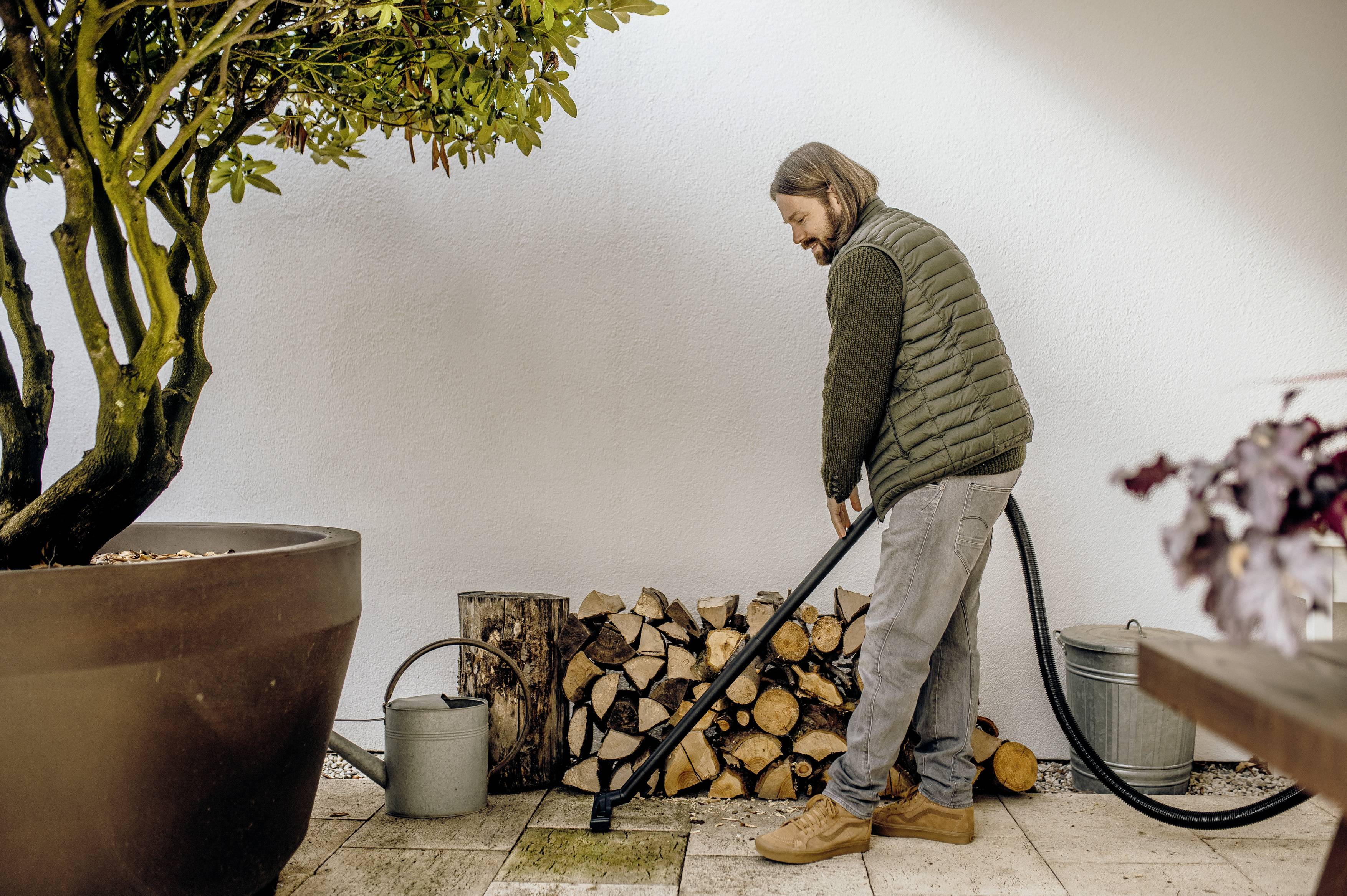 A person is watering plants with a hosepipe in a garden. There are stacked logs and watering cans nearby.