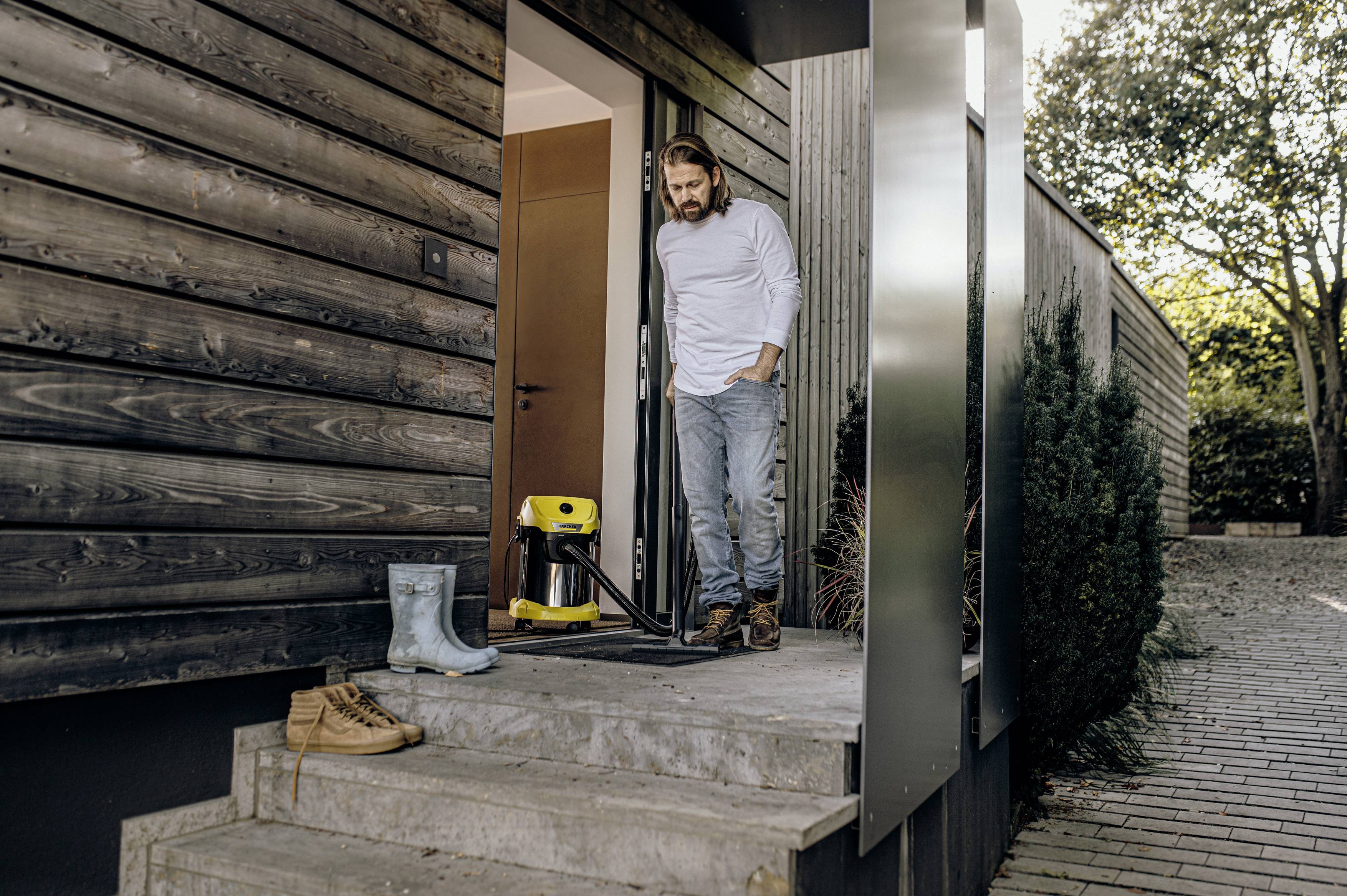 A man is standing in casual clothing on the terrace of a modern wooden house. Next to him is a yellow pressure washer.