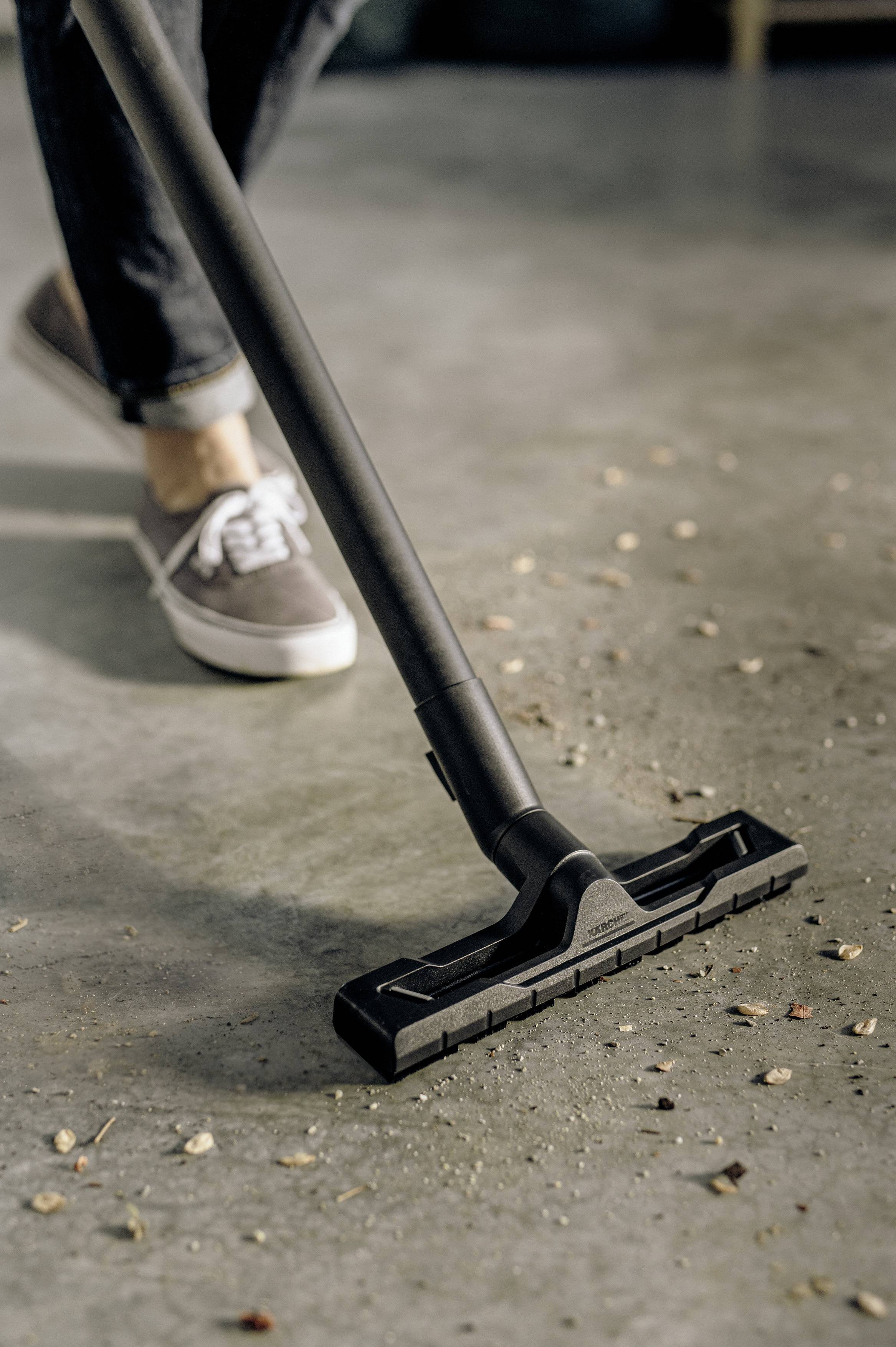 A person is vacuuming leaves and dirt on a grey floor using a vacuum cleaner, wearing casual grey trainers and jeans.