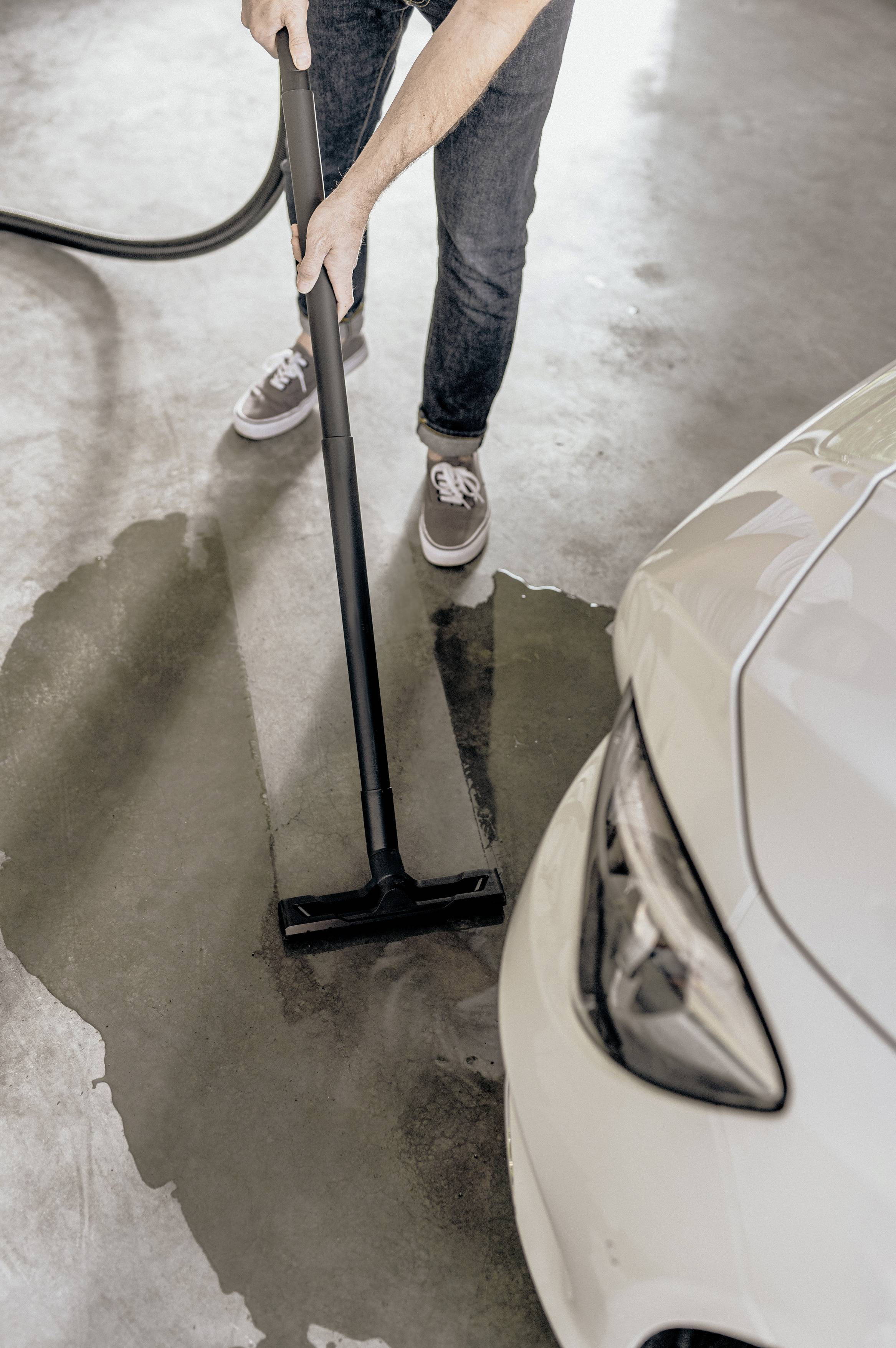 A person is using a wet vacuum cleaner to suck up a puddle of water near a white car in a garage.