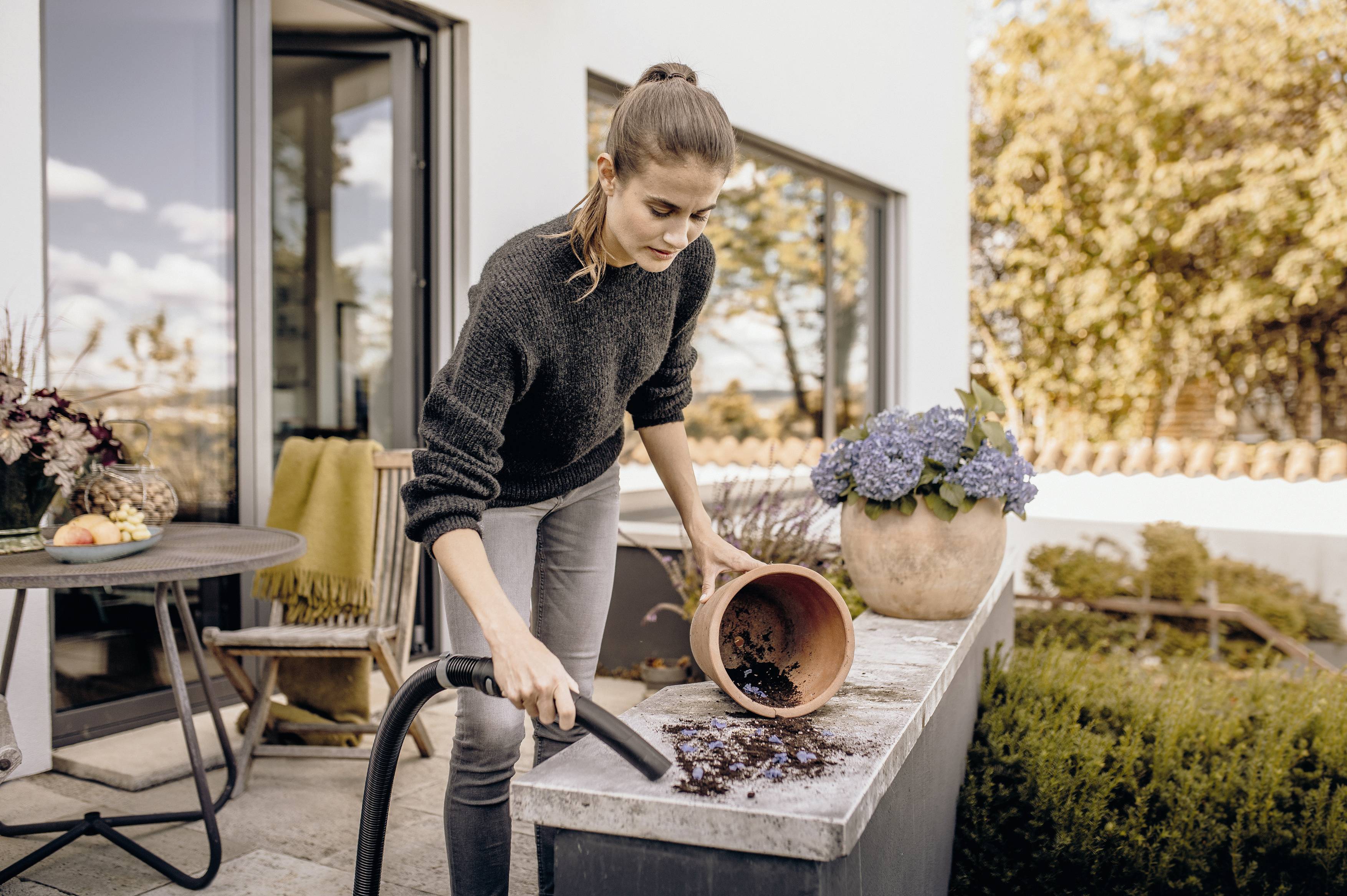 A woman is vacuuming potting soil on an outdoor table. Flowers and a house can be seen in the background.