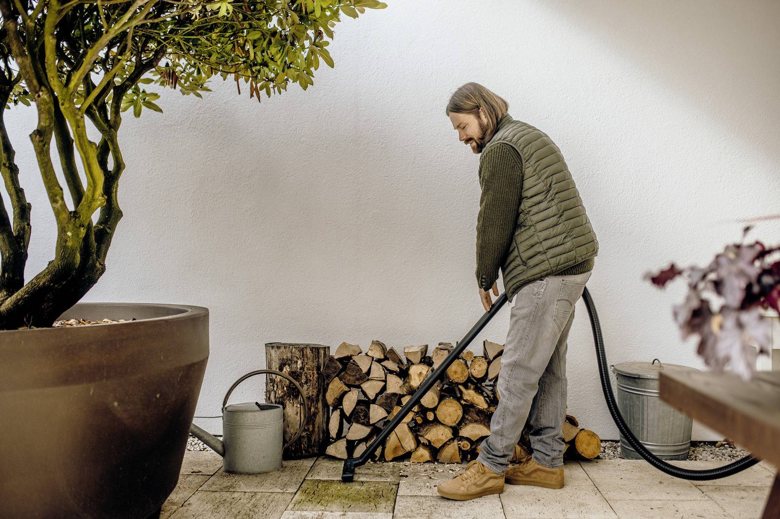 A person with a hose is watering potted plants on a patio. There are logs stacked against the wall, and a watering can is nearby.