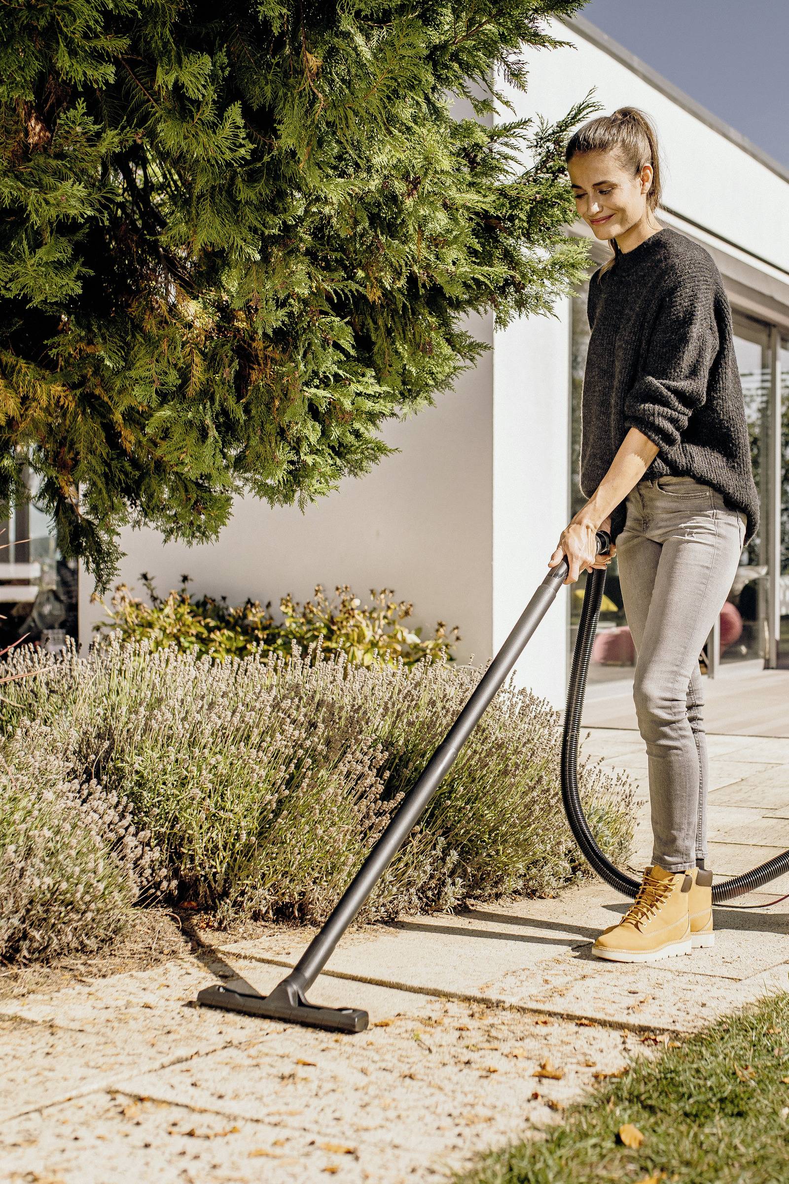 Person vacuuming a garden pathway next to a lavender bush, wearing a dark sweater and light jeans, with a modern building in the background.