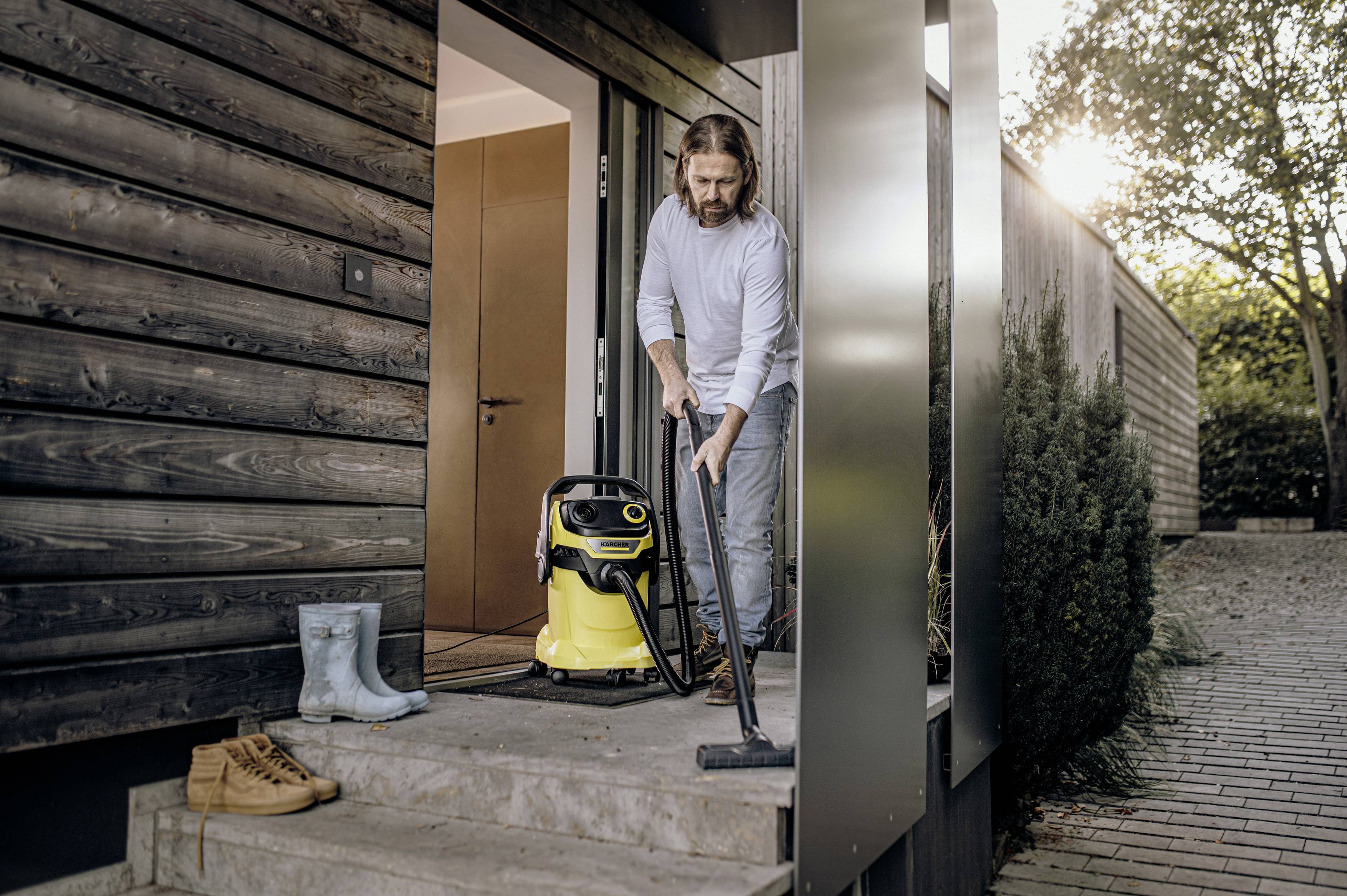 A man is using a yellow vacuum cleaner on the entrance steps of a modern house. Boots are standing beside the door.