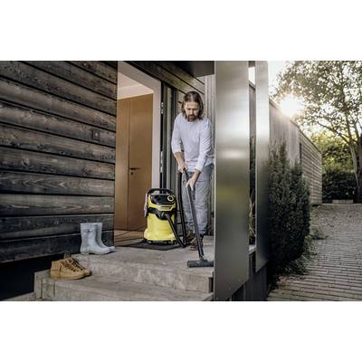 A man is using a yellow vacuum cleaner on the entrance steps of a modern house. Boots are standing beside the door.