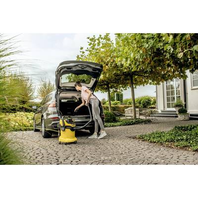 A person is vacuuming the boot of a car with a yellow hoover on a tree-lined, paved driveway.